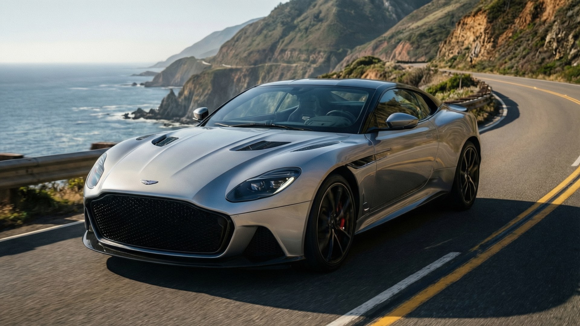 Aston Martin Coastal 壁紙 — 一輛銀色 Aston Martin DBS Superleggera 沿著海岸公路飛馳，背景是懸崖和海洋。