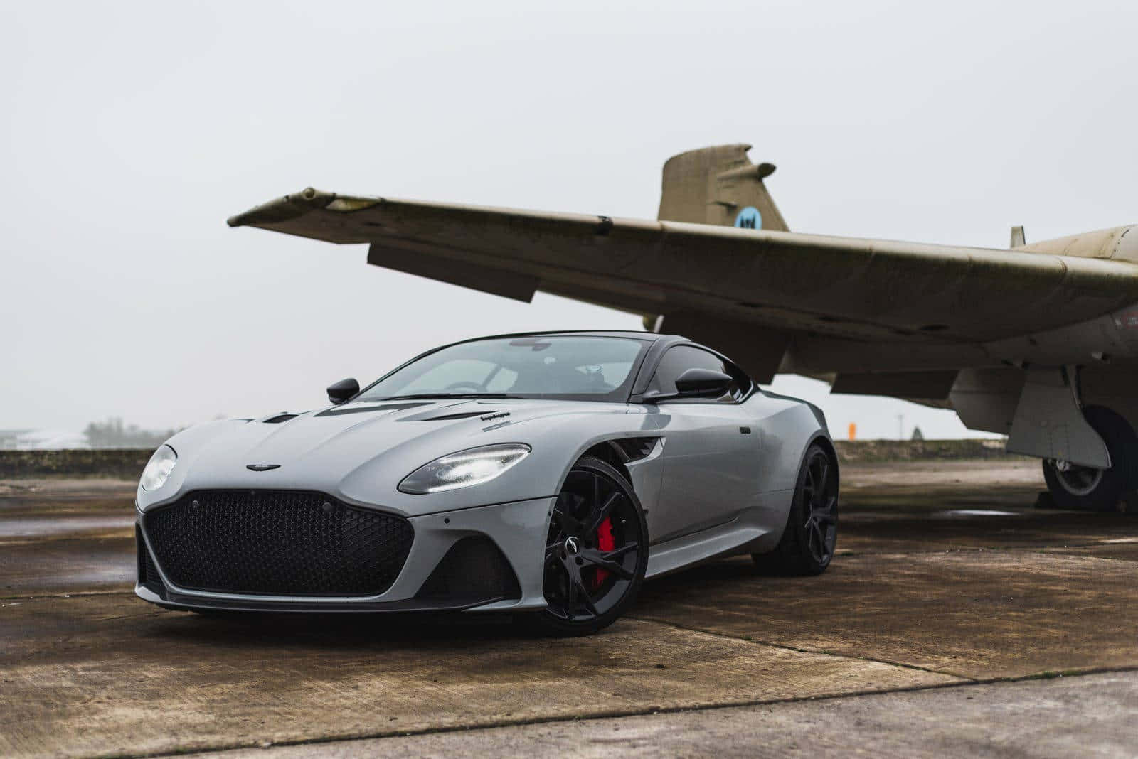 Aston Martin Dbs Superleggera 桌布 壁紙 — 一架時尚的銀色Aston Martin跑車停放在濕滑的停機坪上，旁邊是一架大型復古軍用飛機，天空多雲。