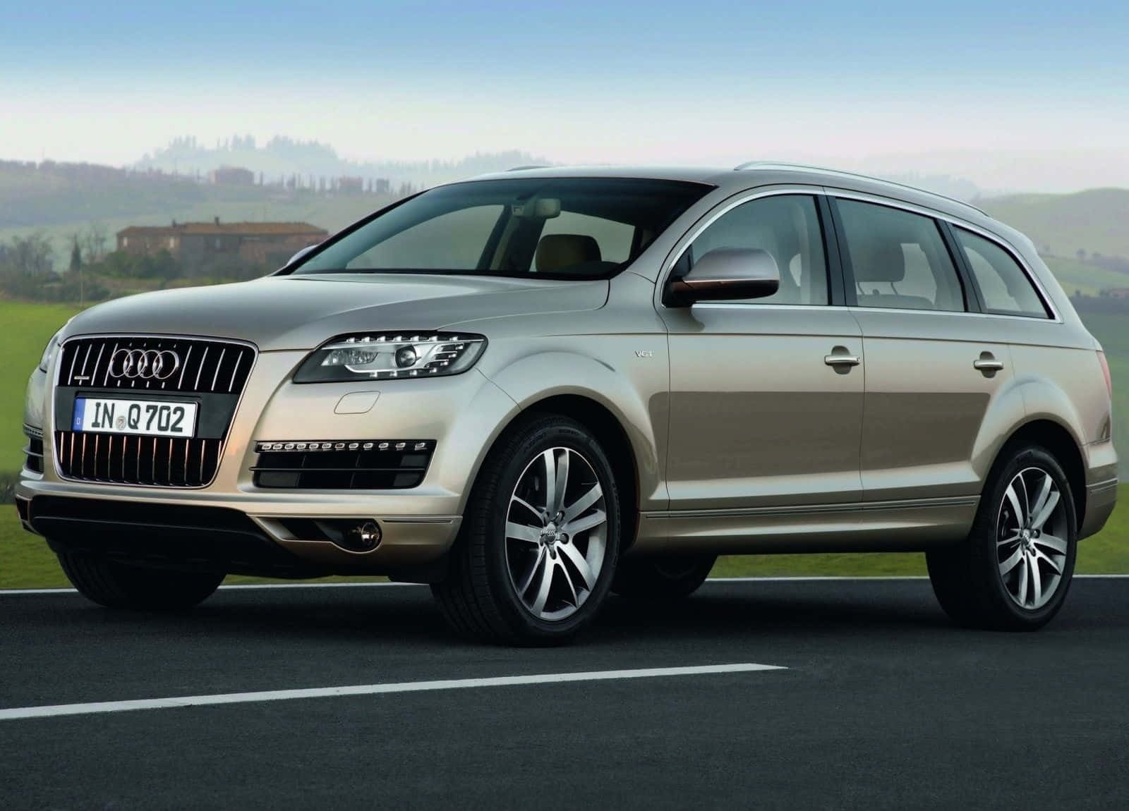 Audi Q7 桌布 壁紙 — 一輛時尚的銀色奧迪Q7 SUV停泊在柏油路上，周圍是連綿起伏的綠色山丘，遠處有一座農舍，天空清澈湛藍。