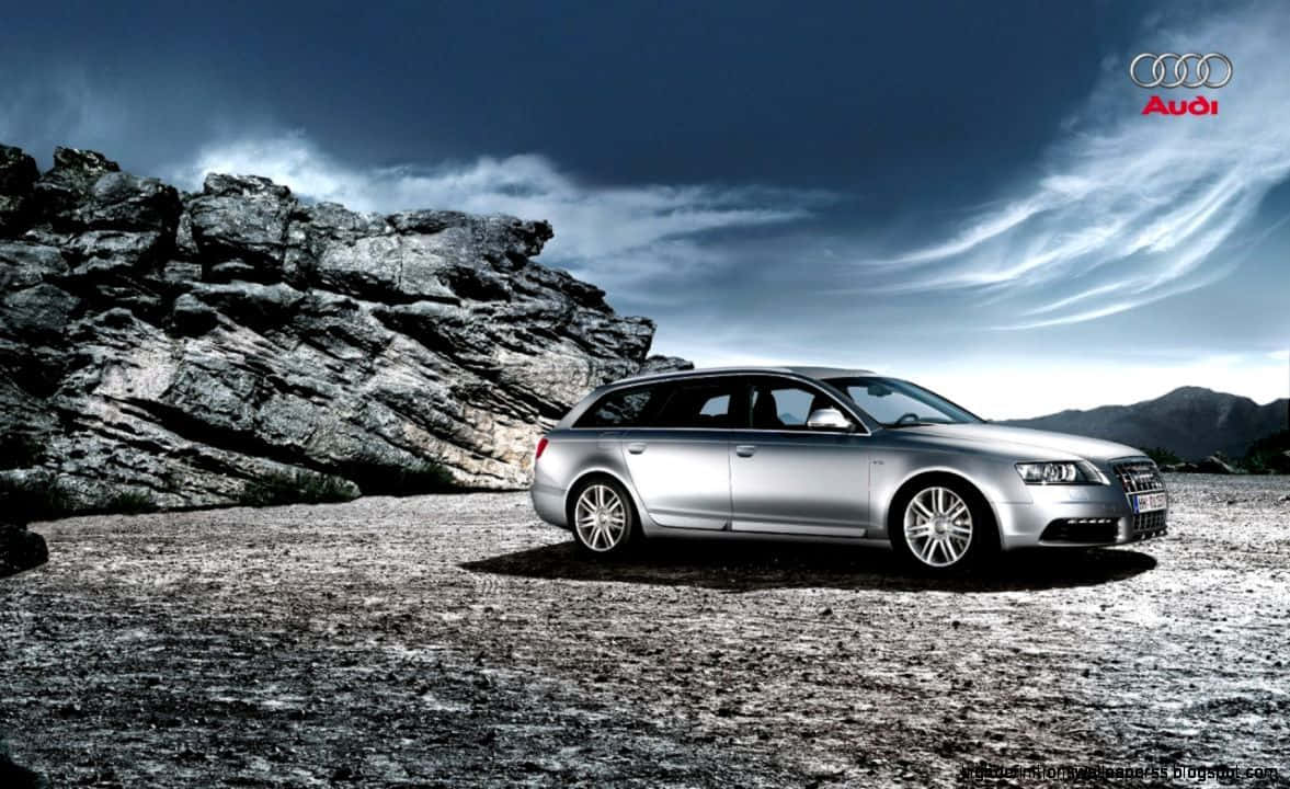 Audi S6 桌布 壁紙 — 一輛時尚的銀色奧迪旅行車停在碎石上，旁邊是堅固的岩石形成，背景是戲劇性的多雲天空。