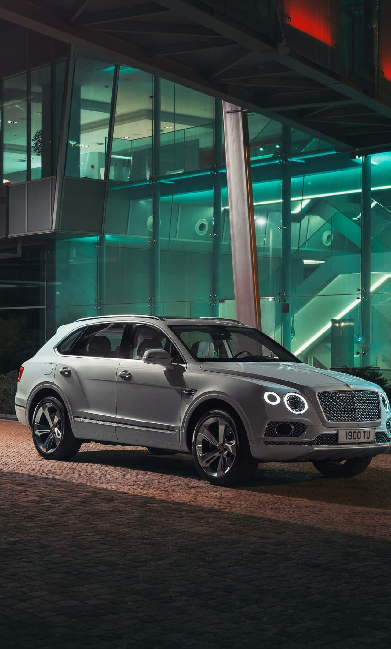 Bentley Bentayga 桌布 壁紙 — 一輛銀色賓利Bentayga SUV在夜間停泊在現代玻璃建築前，在霓虹燈下閃閃發光。