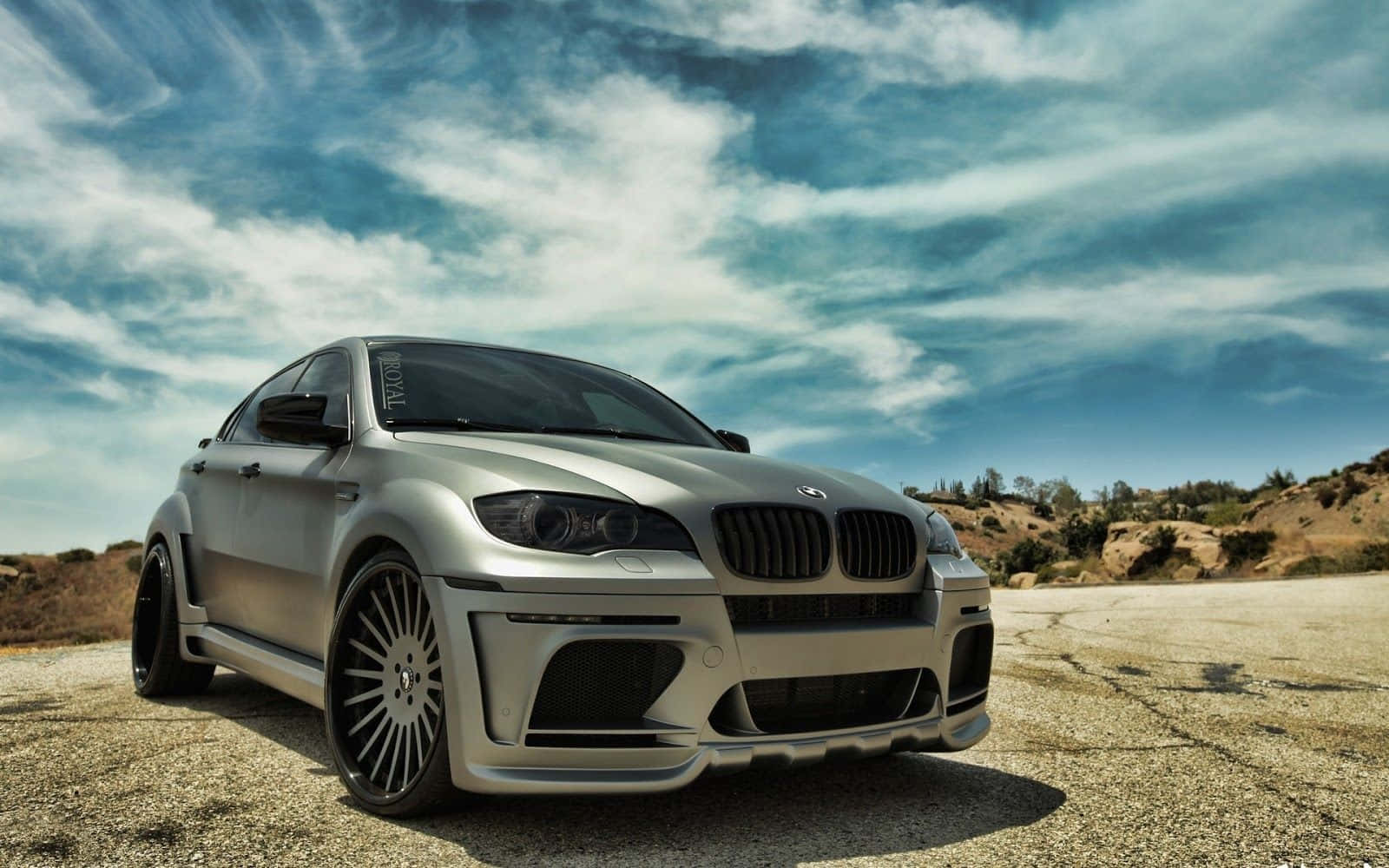 Bmw X6 桌布 壁紙 — 一輛銀色BMW M6雙門跑車配備客製化寬體套件，停在裂開的柏油路面上，天空多雲而戲劇化。