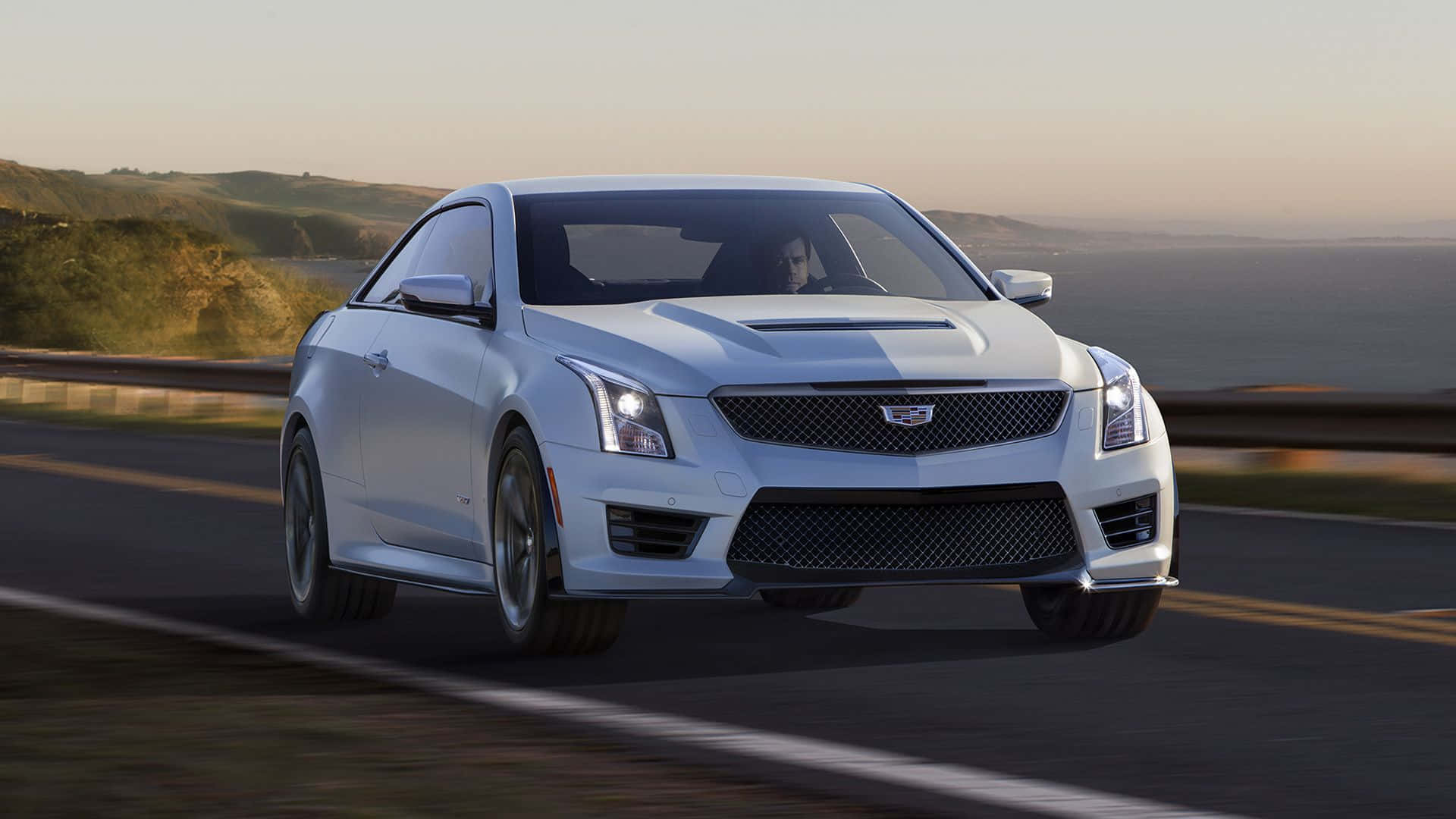 Cadillac Ats 桌布 壁紙 — 一輛時尚的銀色Cadillac ATS-V轎跑車在日落時分沿著海岸高速公路飛馳，背景是模糊的山丘和海洋。