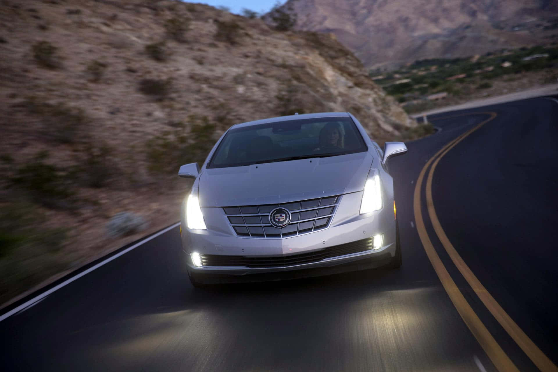 Cadillac Elr 桌布 壁紙 — 一輛銀色Cadillac房車在黃昏時分沿著蜿蜒的山路疾馳，車頭燈閃爍。