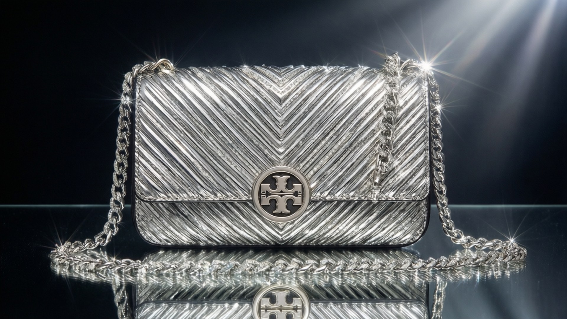 Silver Kira Clutch Sparkle 壁紙 — 銀色人字形圖案手提包，配有鏈條肩帶和標誌獎章，在聚光燈下於反光黑色表面上閃閃發光。