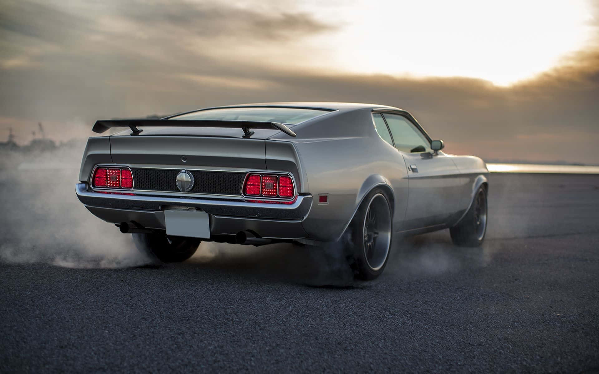 Ford Mustang Mach 1 桌布 壁紙 — 一輛銀色經典肌肉車在金色夕陽天空下甩尾，後輪冒出滾滾濃煙。