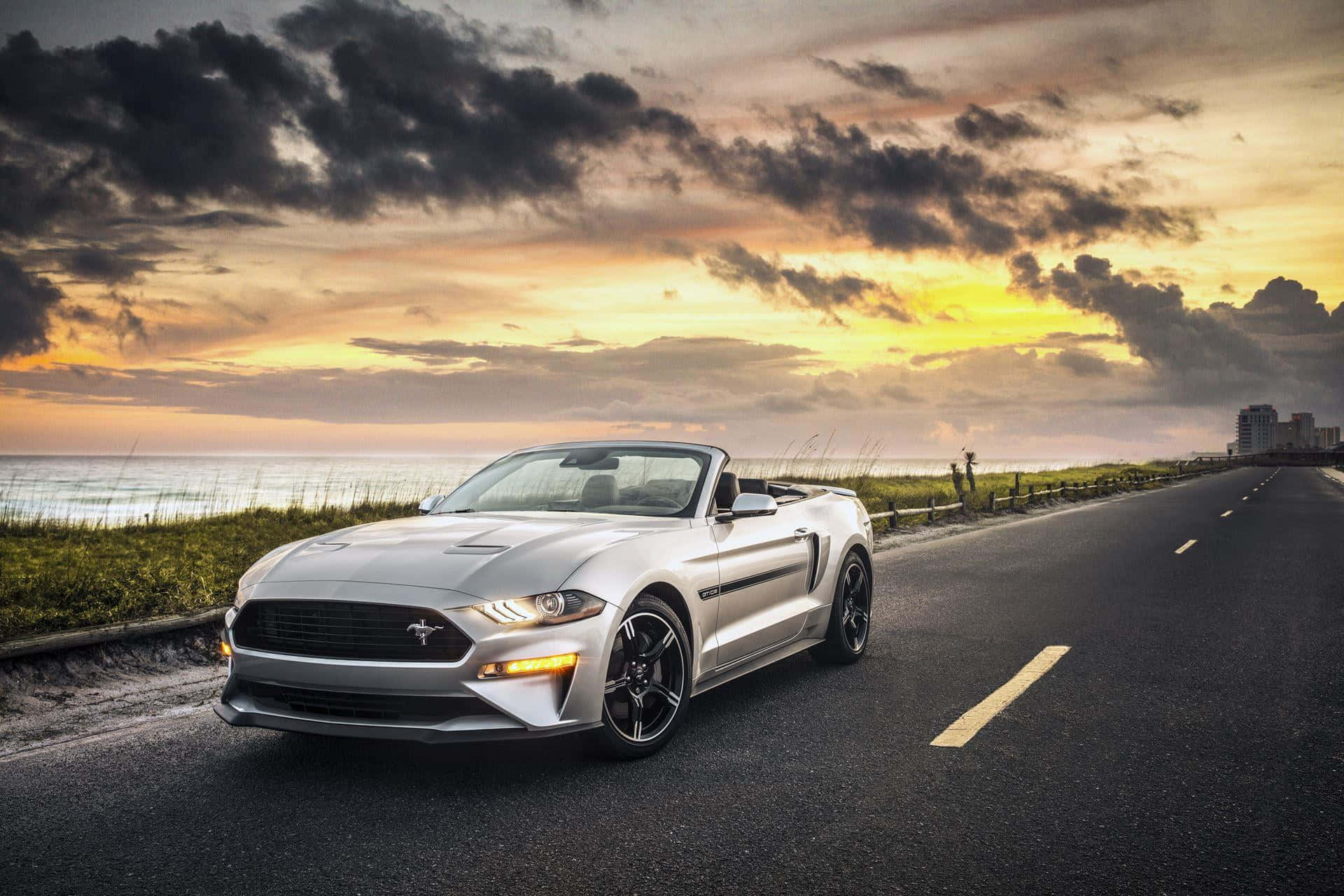 Ford Mustang California Special 桌布 壁紙 — 一輛銀色敞篷跑車在日落時分沿著海岸公路行駛，背景是戲劇性的雲層和海洋。