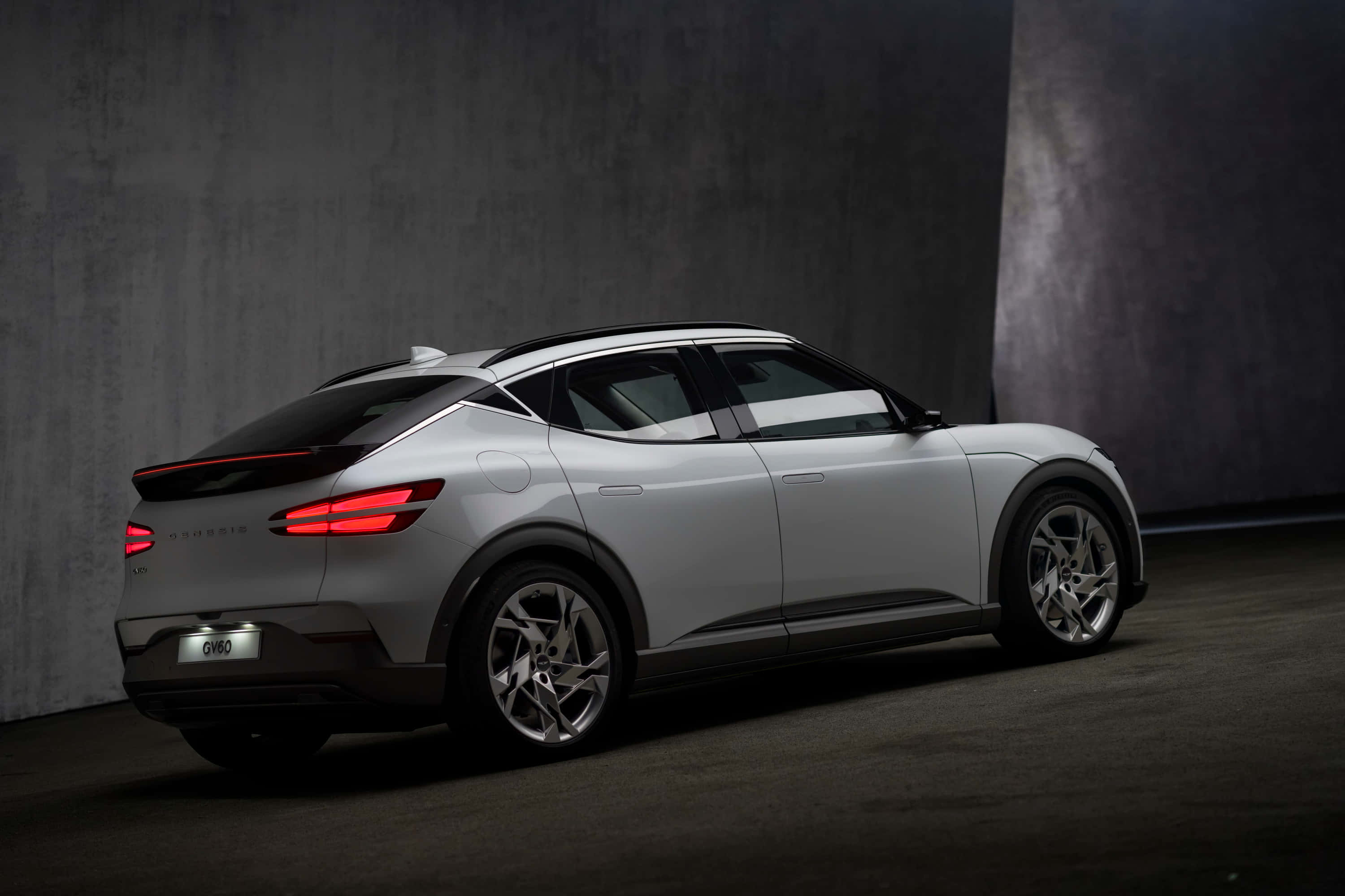 Genesis Gv60 桌布 壁紙 — 一輛時尚的銀色電動SUV，帶有發光的紅色尾燈，在昏暗的攝影棚中，靠著帶紋理的灰色混凝土牆壁。