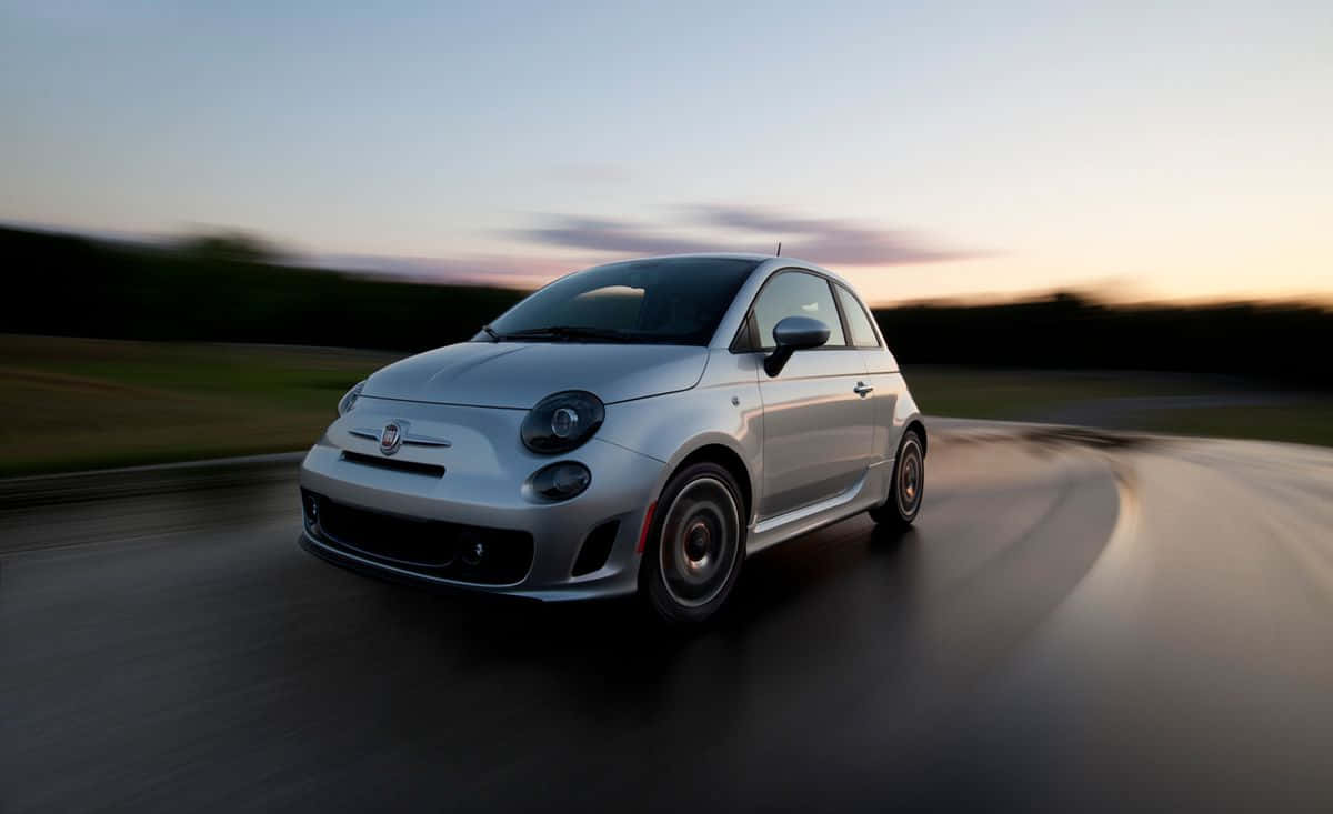 Fiat 500 桌布 壁紙 — 一輛銀色Fiat 500在日落時分沿著濕滑道路行駛，帶有動態模糊和溫暖的天空色調。