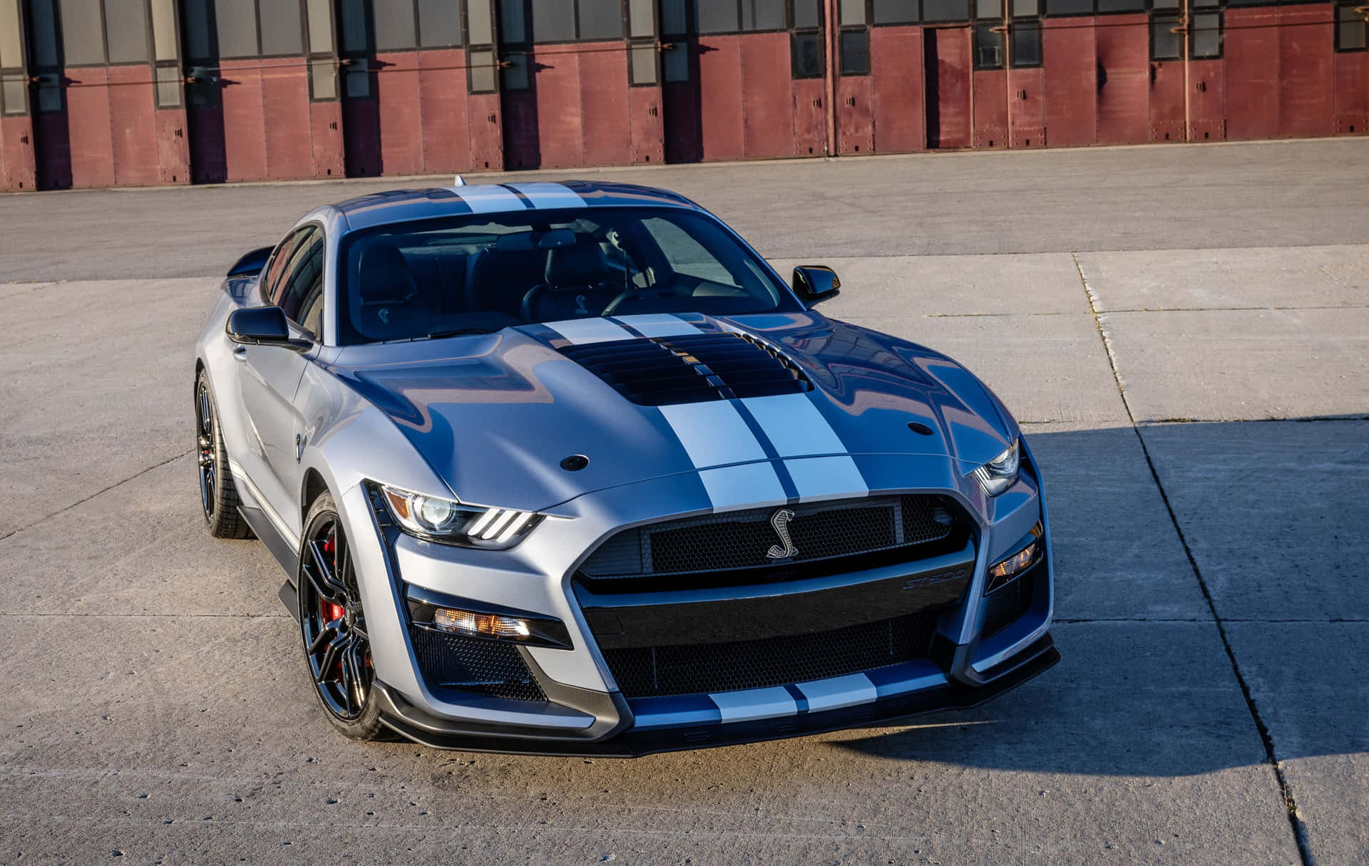  壁紙 — 一輛時尚的銀色福特野馬Shelby GT350，帶有醒目的藍色賽車條紋，停在紅色工業建築前的混凝土上。
