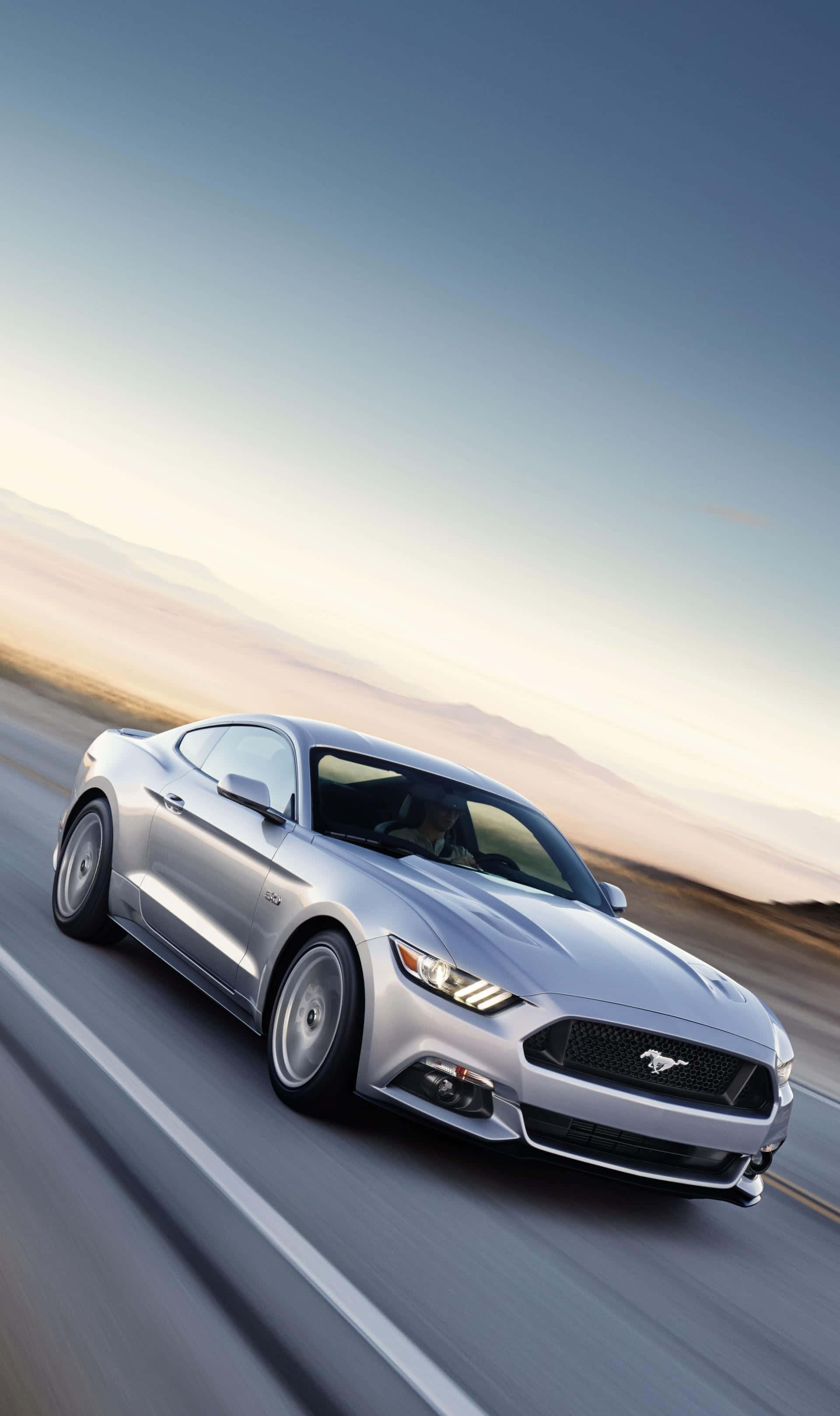 Ford Mustang California Special 桌布 壁紙 — 一輛銀色福特野馬在柔和的金色日落天空下沿著沙漠高速公路飛馳。