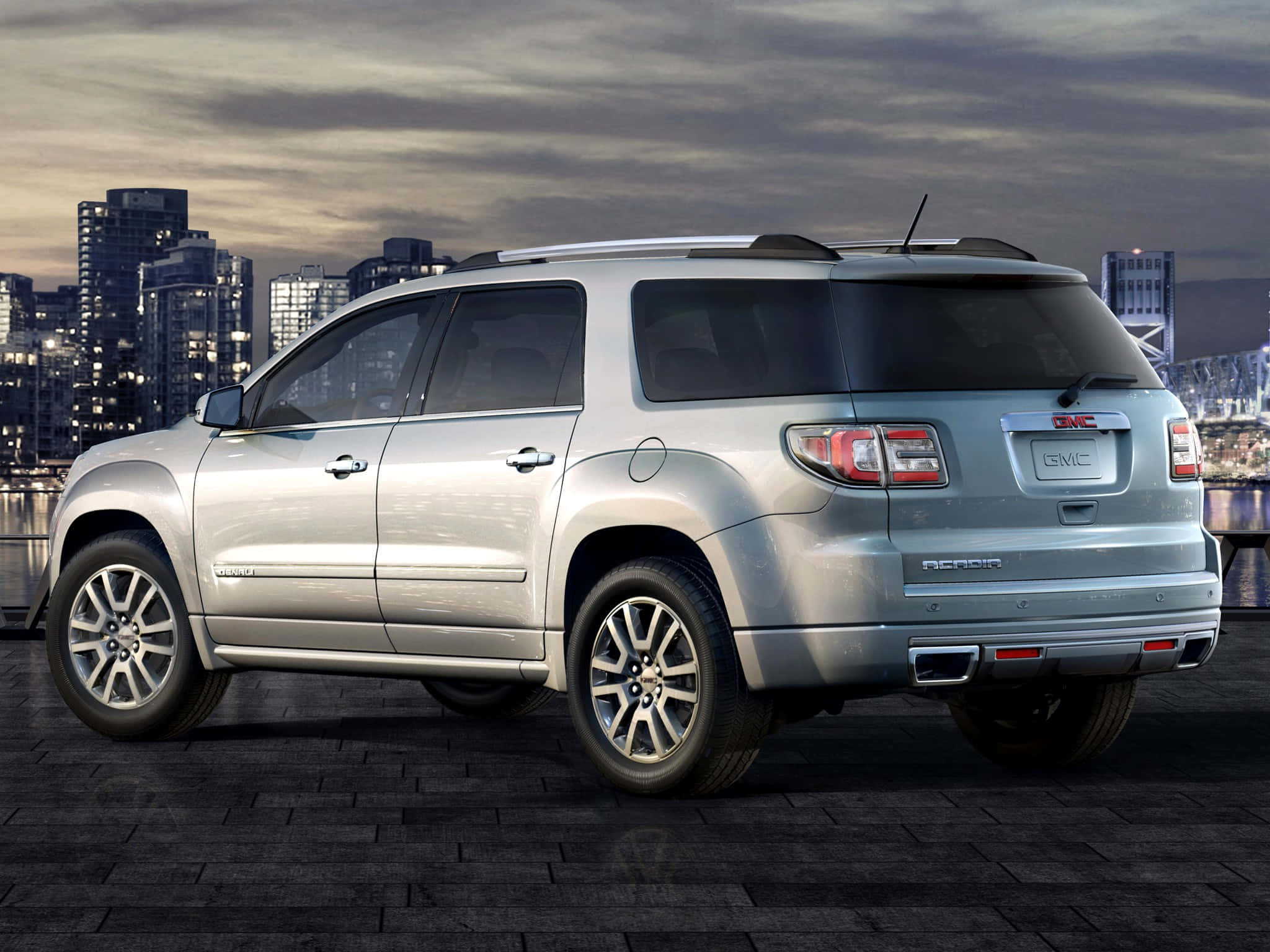 Gmc Acadia 桌布 壁紙 — 一輛銀色GMC Acadia SUV停泊在深色磚砌海濱，背後的城市天際線在黃昏中閃耀。