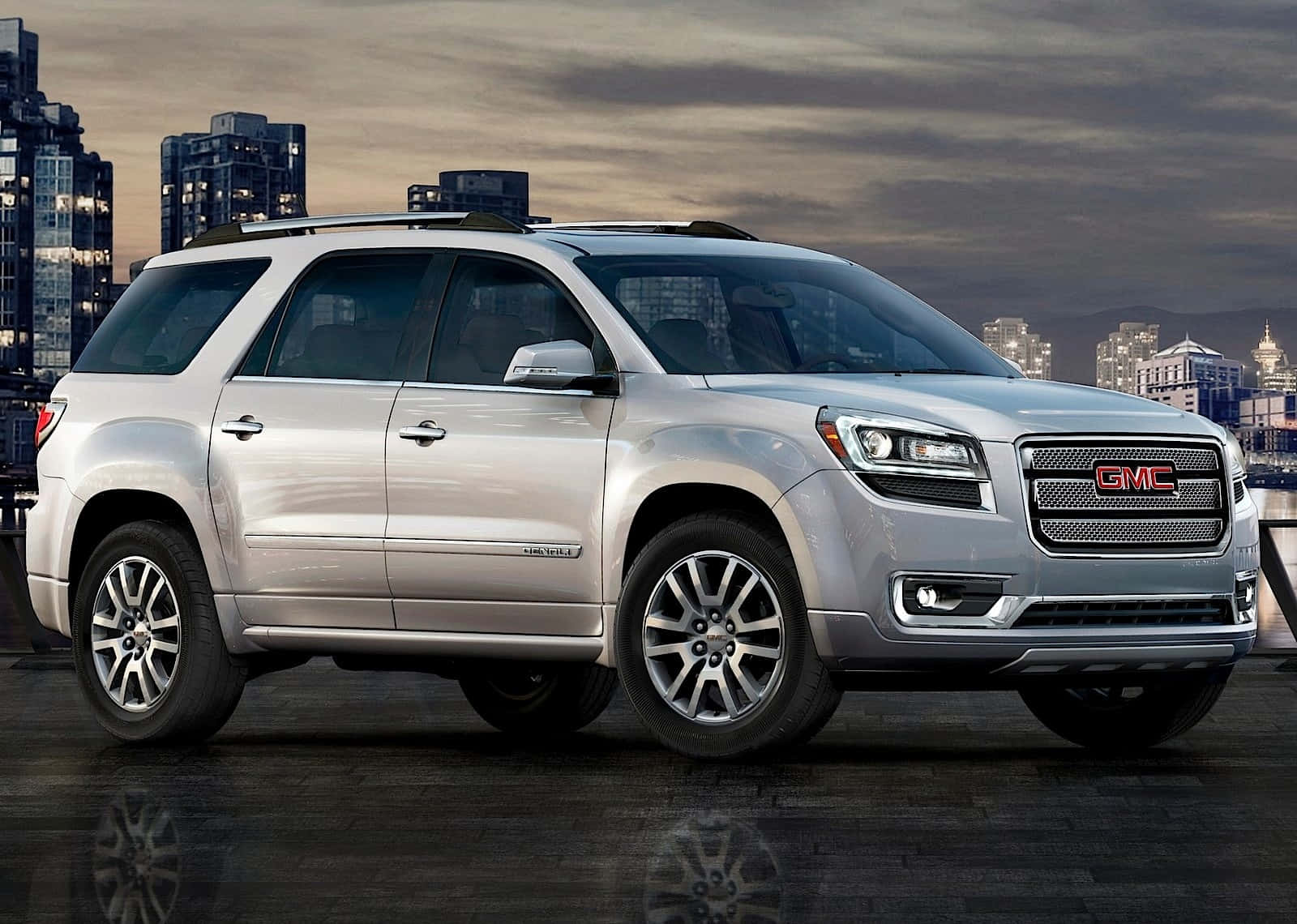 Gmc Acadia 桌布 壁紙 — 一輛銀色GMC Acadia SUV停泊在黃昏時濕潤的城市屋頂上，背後城市燈光閃爍。