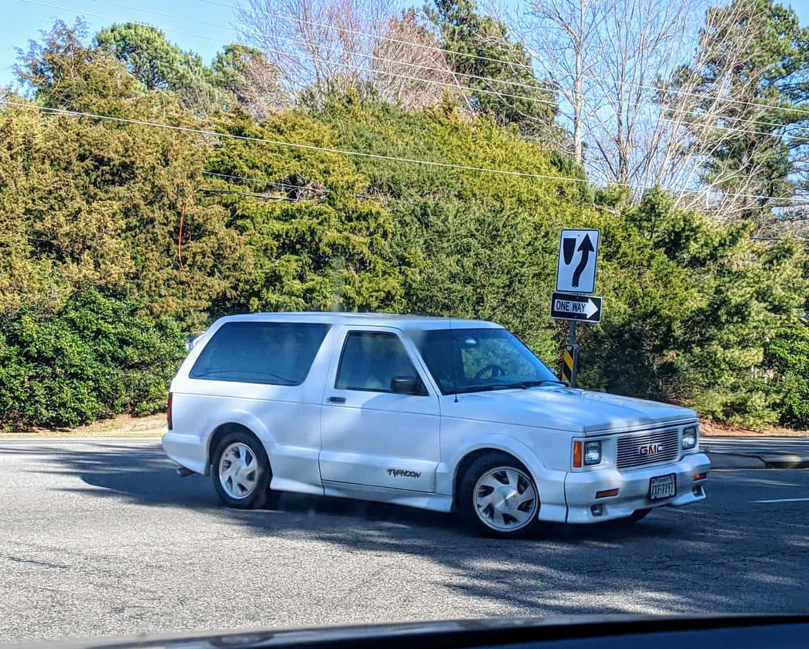 Gmc Typhoon 桌布 壁紙 — 一輛銀色GMC Typhoon SUV在陽光明媚的道路上行駛，道路兩旁是茂密的綠色灌木和光禿禿的樹木，頭頂是清澈的藍天。