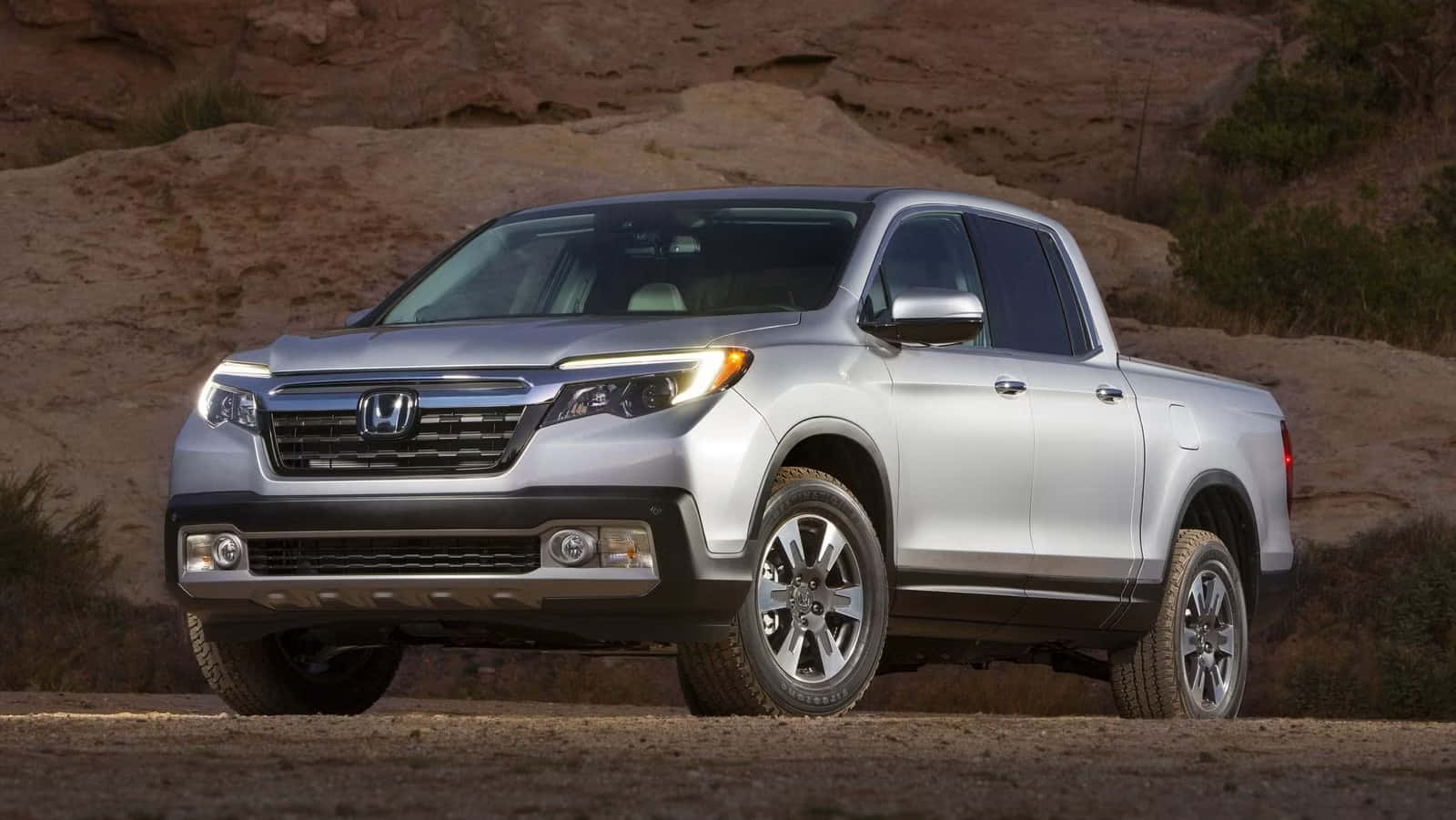 Honda Ridgeline 桌布 壁紙 — 一輛銀色本田 Ridgeline 貨卡停泊在塵土飛揚的沙漠道路上，背景是岩石地形和稀疏的綠色植物。