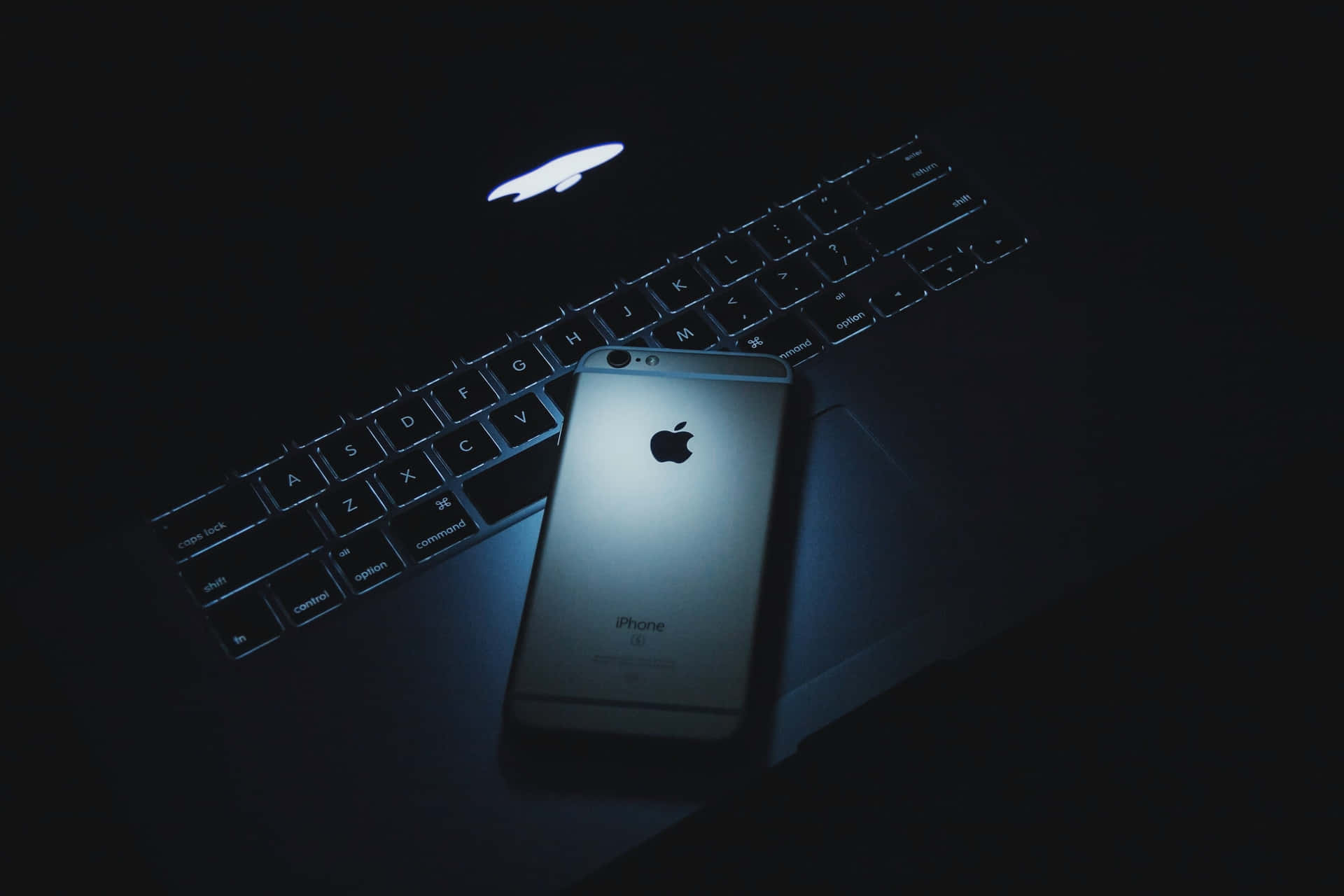 銀色iPhone在筆記型電腦鍵盤上，昏暗藍光 — Macbook Pro 壁紙