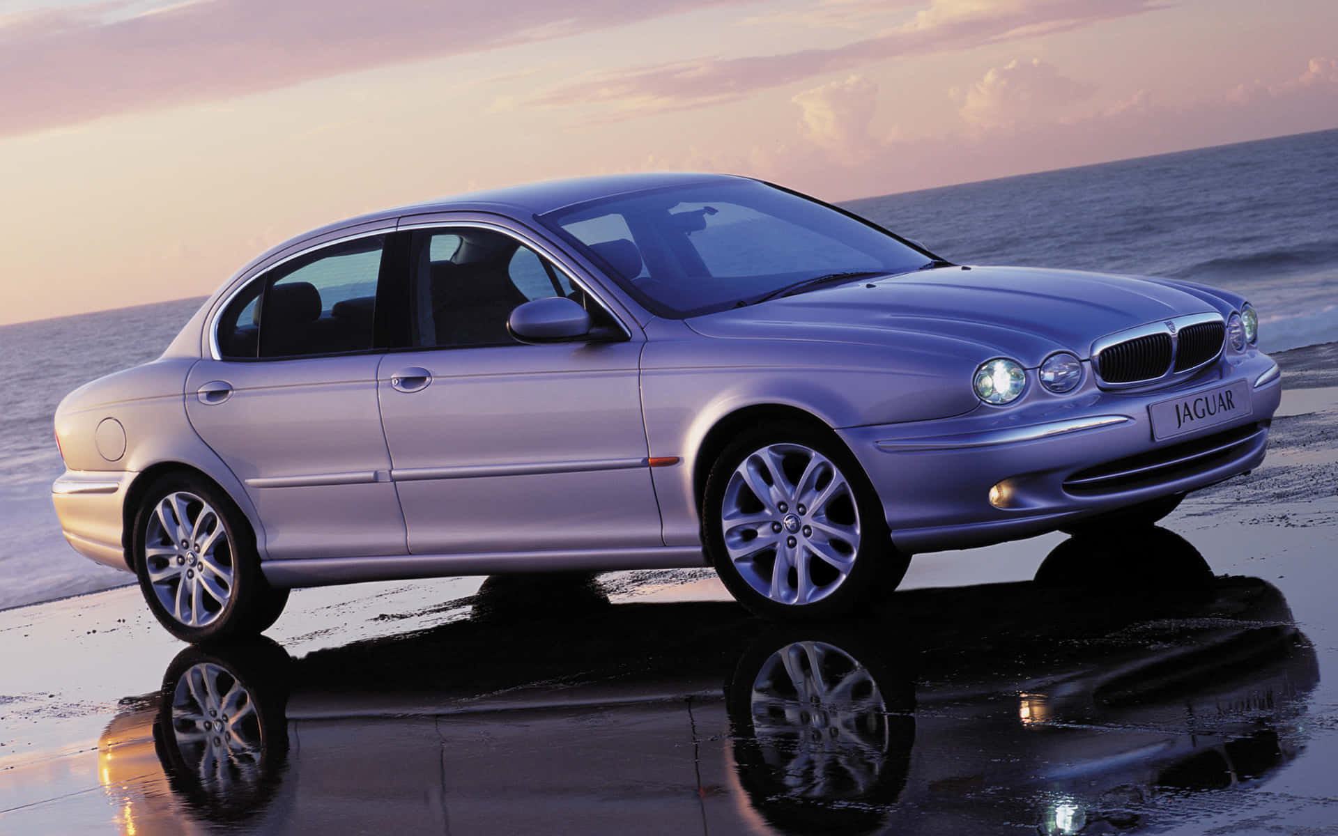 Jaguar X-type 桌布 壁紙 — 一輛銀色積架轎車在日落時分在濕沙上閃閃發光，映照著海浪和天空。