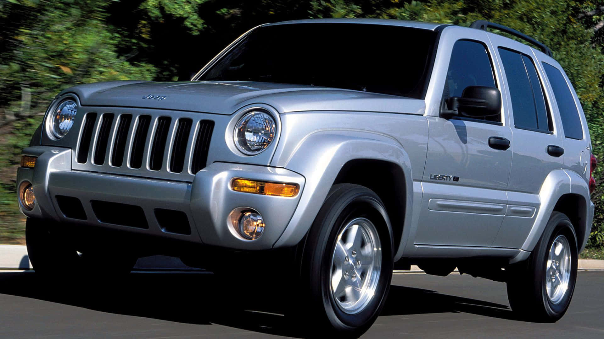 Jeep Liberty 桌布 壁紙 — 一輛銀色Jeep Liberty SUV在道路上行駛，背景有模糊的綠色樹木。