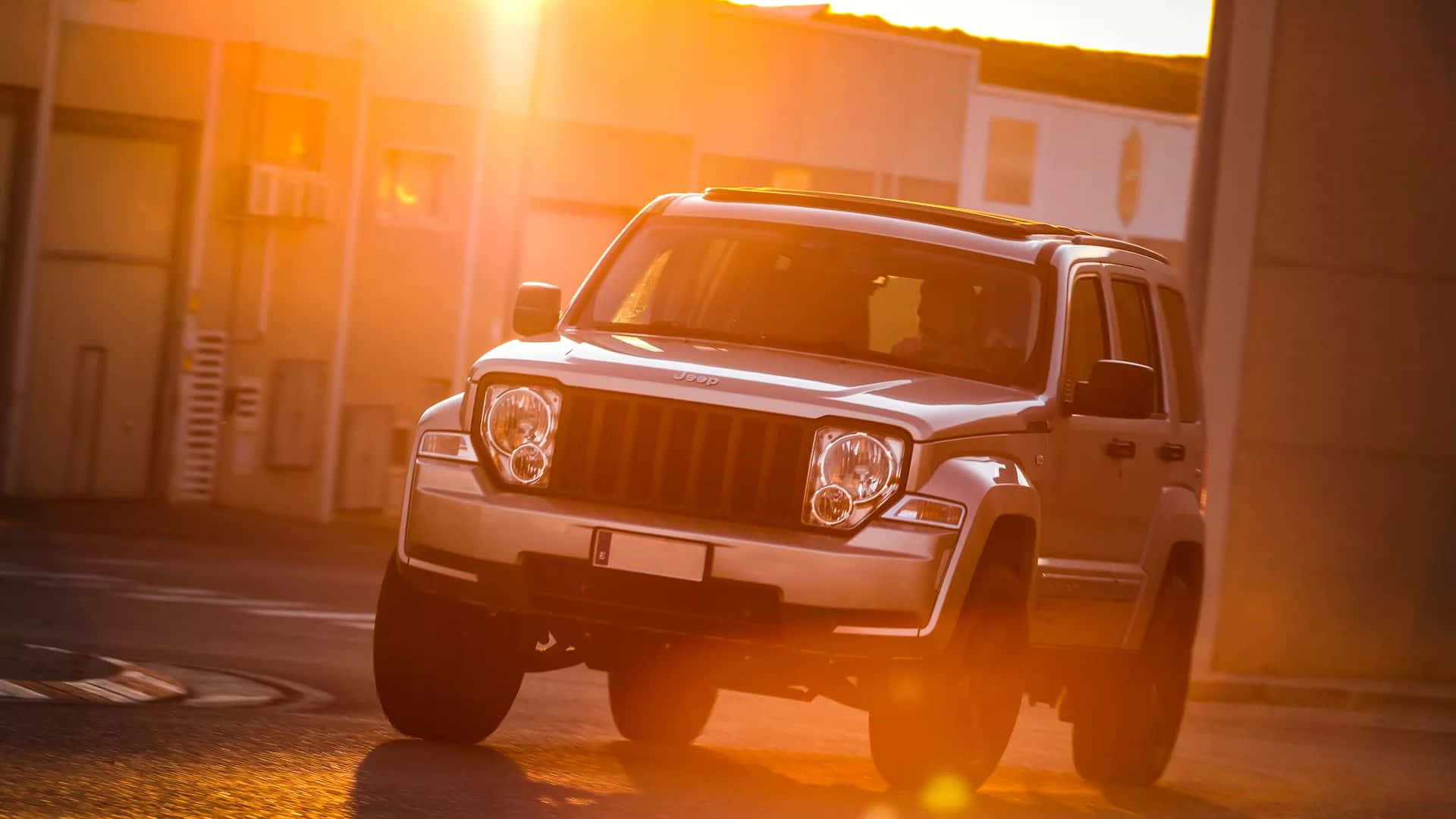 Jeep Liberty 桌布 壁紙 — 一輛銀色Jeep Wrangler停放在都市空地，沐浴在帶有鏡頭光暈的金色夕陽光中。