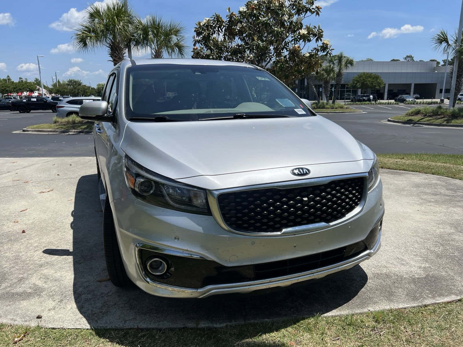 Kia Sedona 桌布 壁紙 — 一輛銀色Kia SUV停放在混凝土墊上，背景是明亮的藍天、散落的雲朵和棕櫚樹。