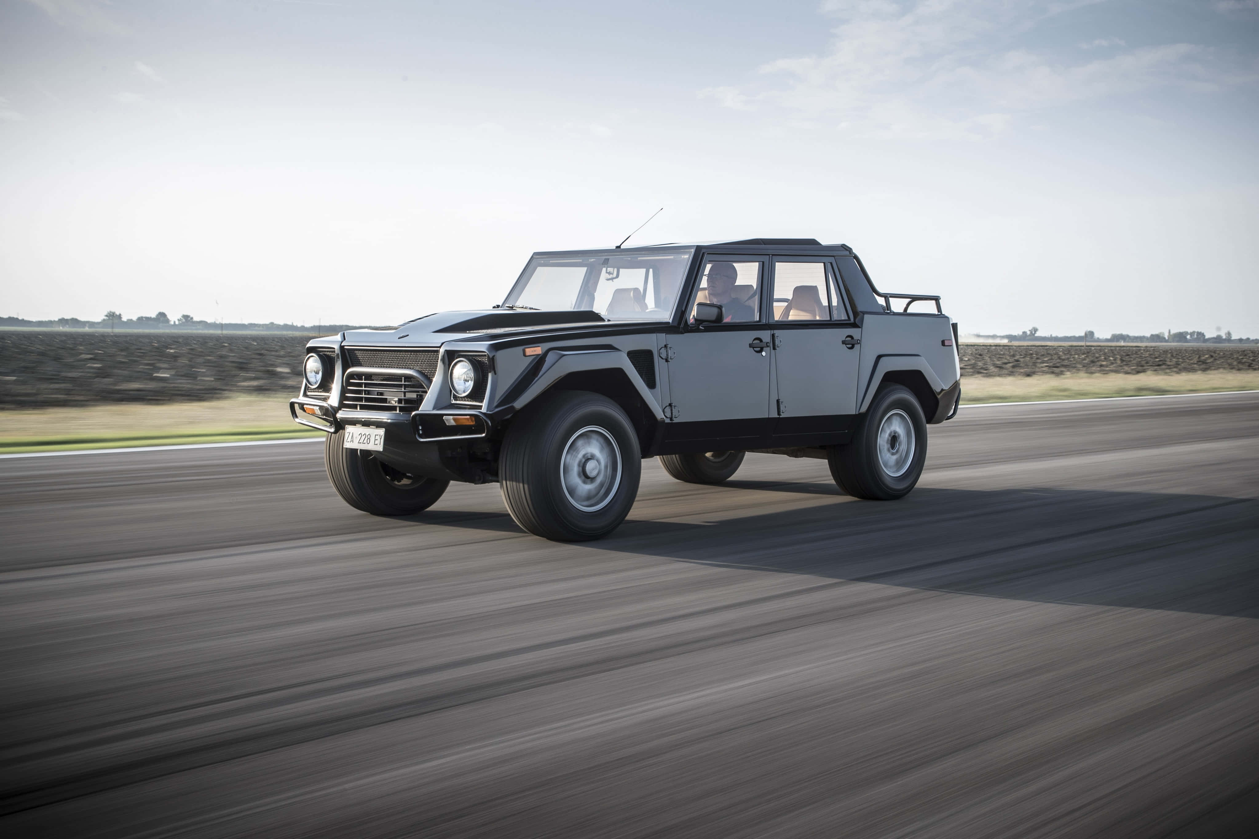 Lamborghini Lm002 桌布 壁紙 — 一輛銀色藍寶堅尼LM002 SUV在陽光照耀的柏油路上奔馳，背景是模糊的動態和開闊的田野。
