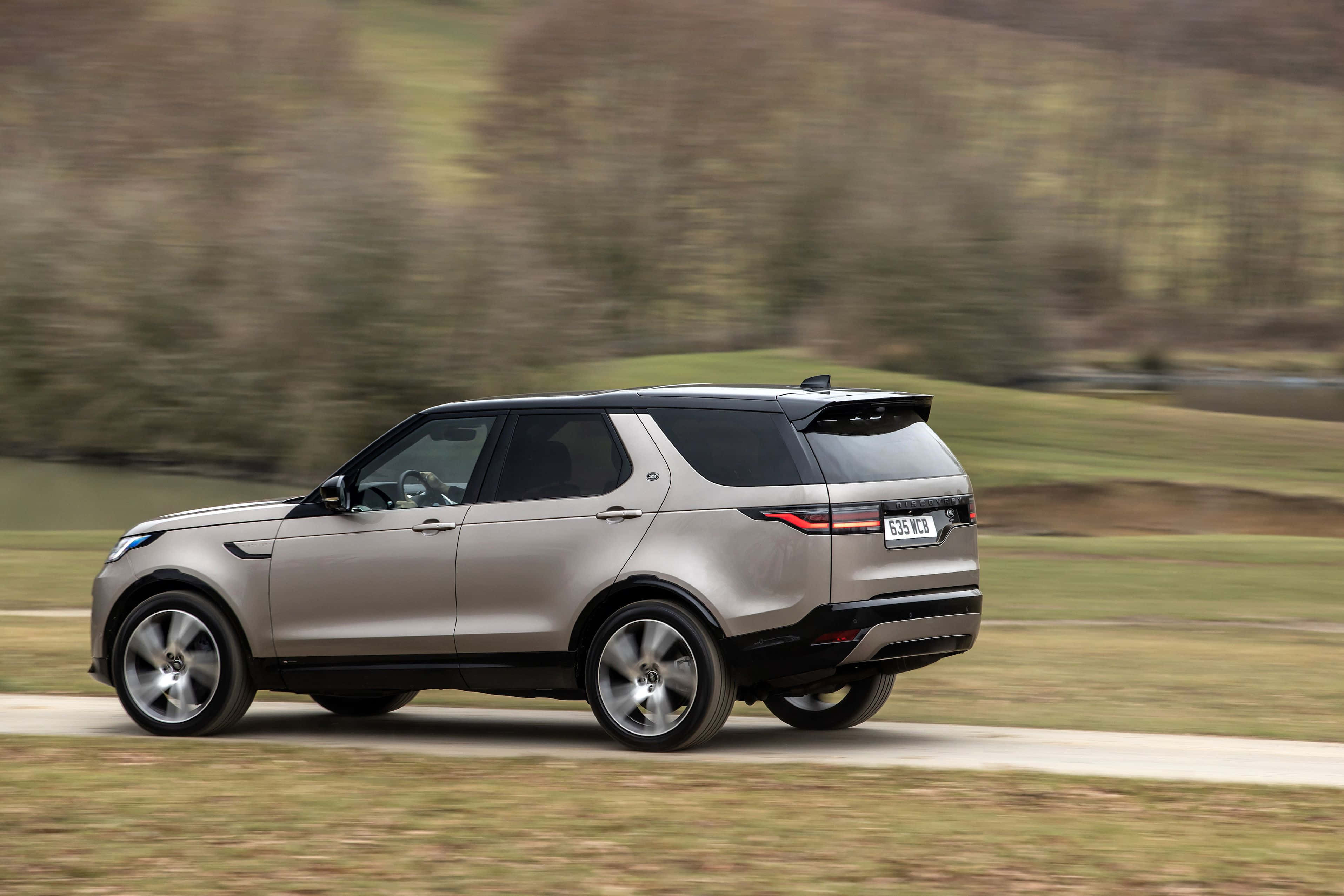 Land Rover Discovery 桌布 壁紙 — 一輛銀色 Land Rover Discovery SUV 在鄉村道路上疾駛，背景是模糊的綠色山丘。