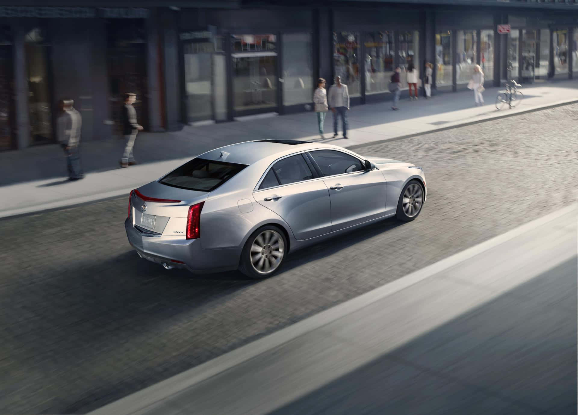 Cadillac Ats 桌布 壁紙 — 一輛銀色豪華轎車在陽光明媚的城市街道上平穩行駛，背景是模糊的行人和商店櫥窗。