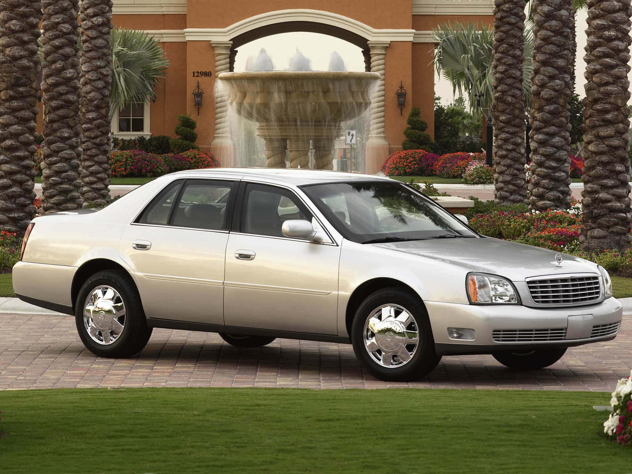 Cadillac Deville 桌布 壁紙 — 一輛銀色豪華轎車停在磚砌車道上，前方有噴泉和棕櫚樹。