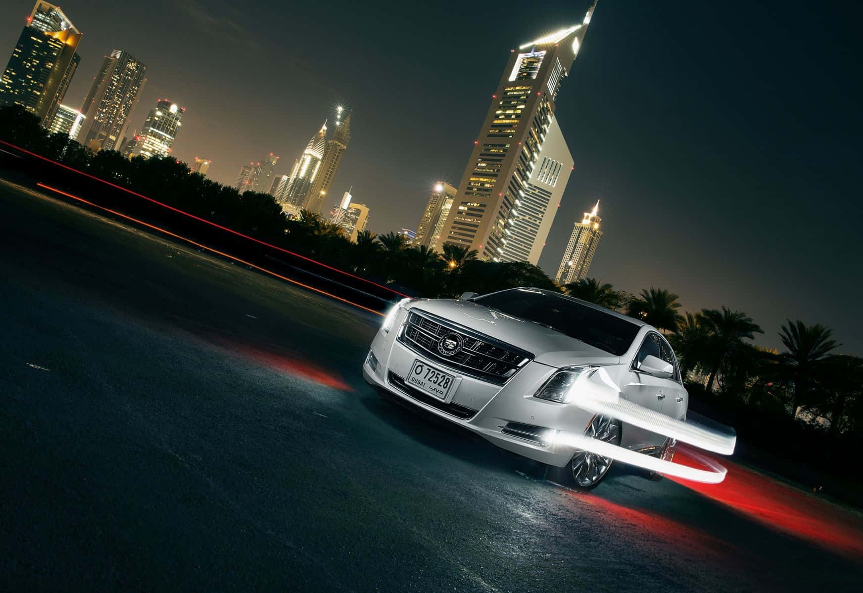 Cadillac Xts 桌布 壁紙 — 一輛銀色豪華轎車在夜間城市景觀中疾馳，身後是發光的摩天大樓和紅色光跡。