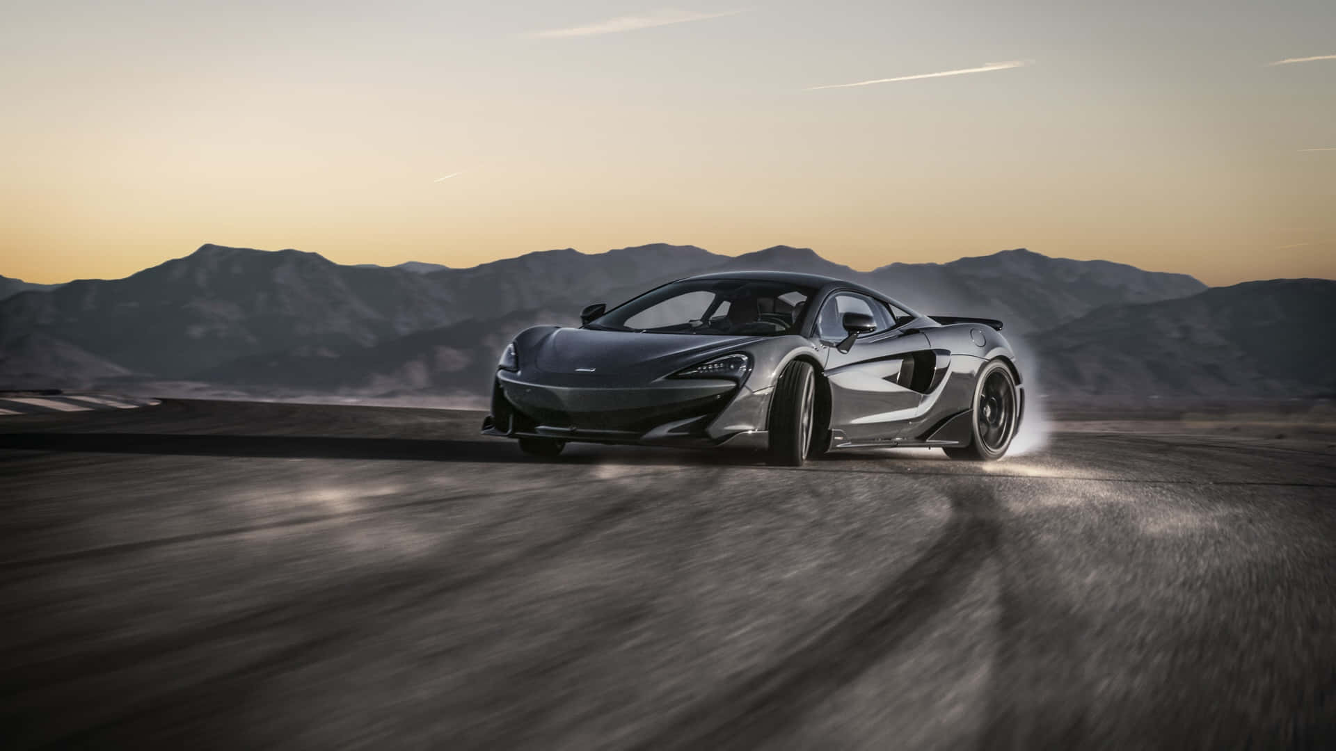 Mclaren 600lt 桌布 壁紙 — 一輛時尚的銀色McLaren跑車在日落時分於沙漠公路上飄移，背景有山脈。