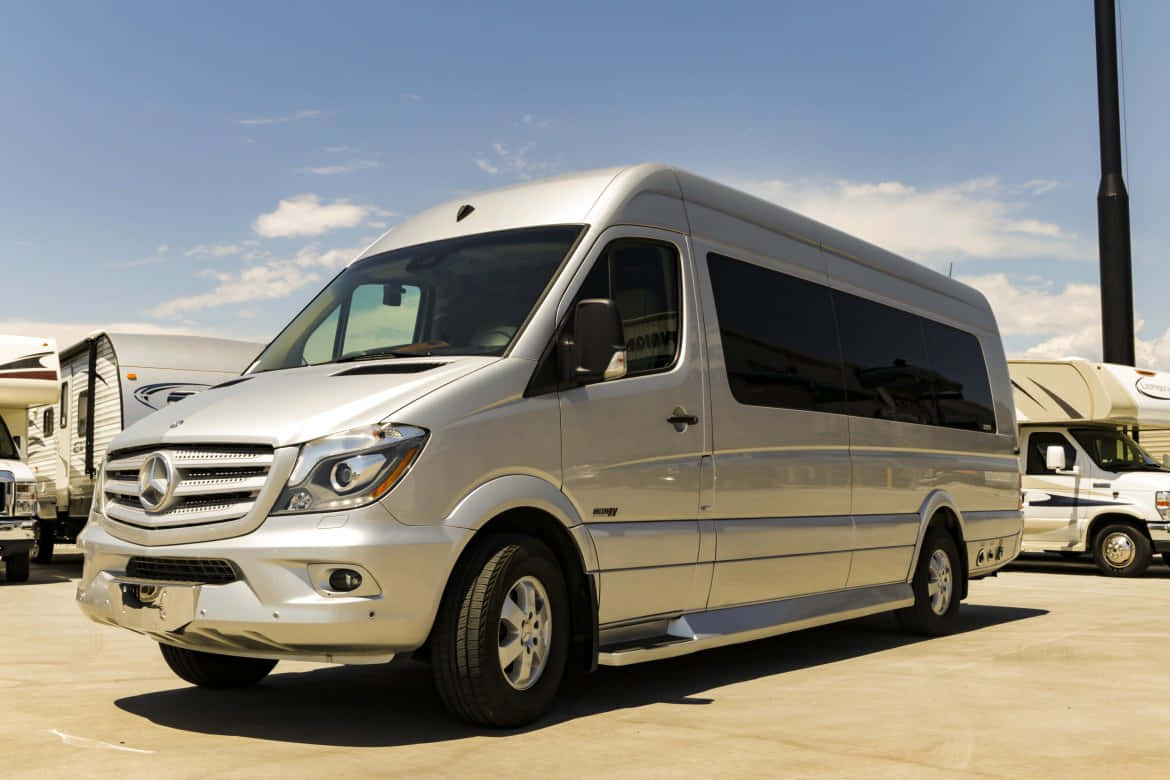 Mercedes Benz Sprinter 桌布 壁紙 — 一架時尚的銀色 Mercedes-Benz Sprinter 客貨車停放在陽光明媚的停車場，背景有露營車，天空蔚藍。