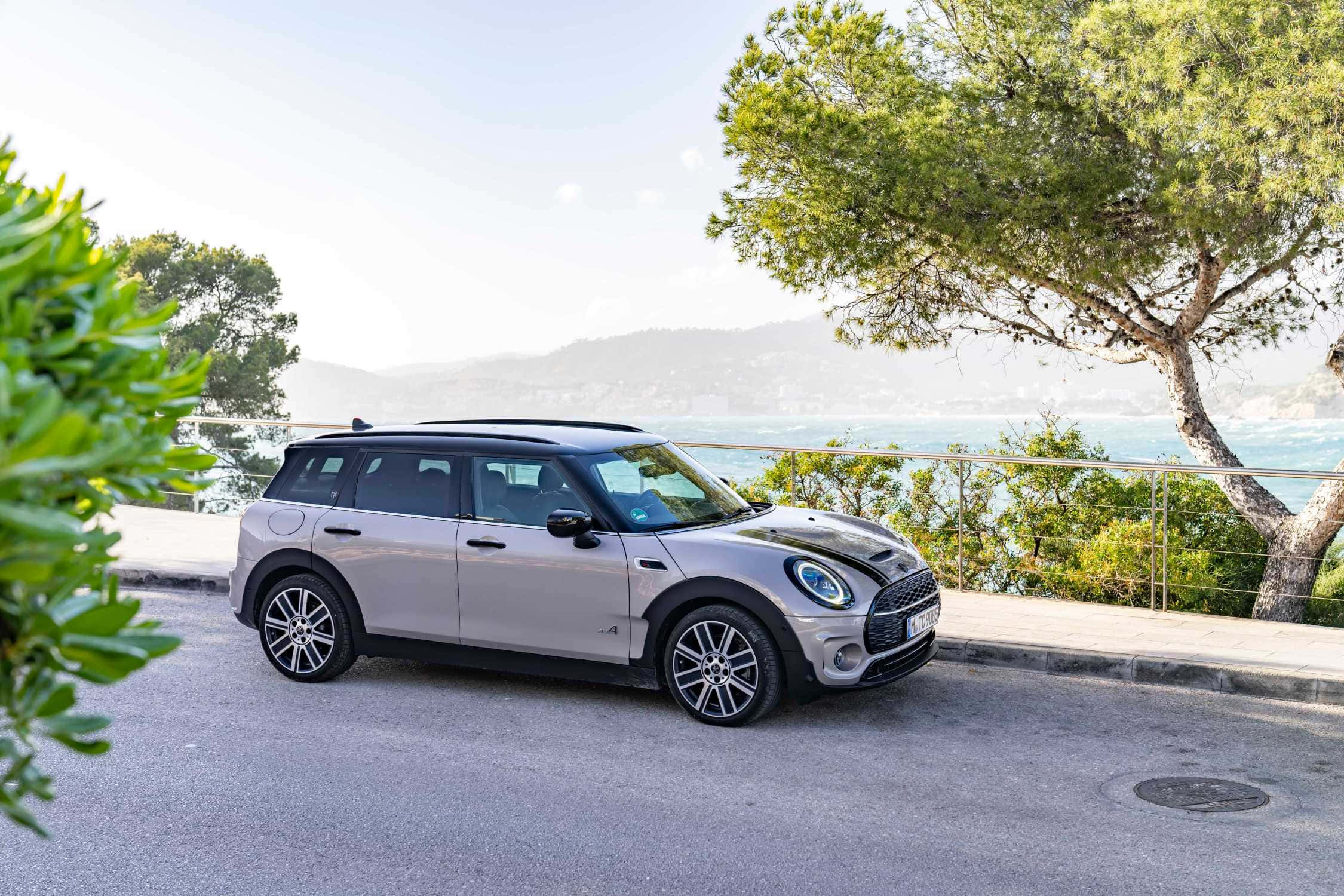 Mini Cooper S Clubman All4 桌布 壁紙 — 一輛銀色MINI Countryman停泊在沿海道路上，周圍有茂密的綠樹和遠處的山巒海岸線，在明亮的日光下。