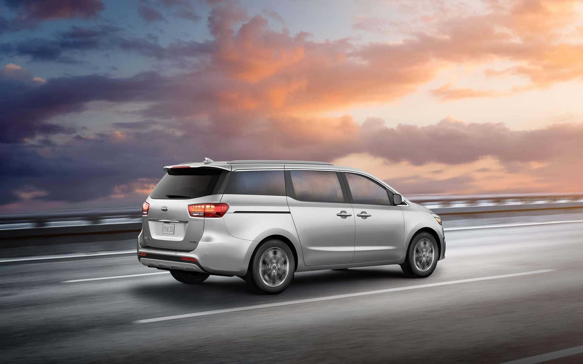 Kia Sedona 桌布 壁紙 — 一輛銀色小型貨車在高速公路上行駛，天空是壯麗的日落，帶有橙色和紫色的雲彩。