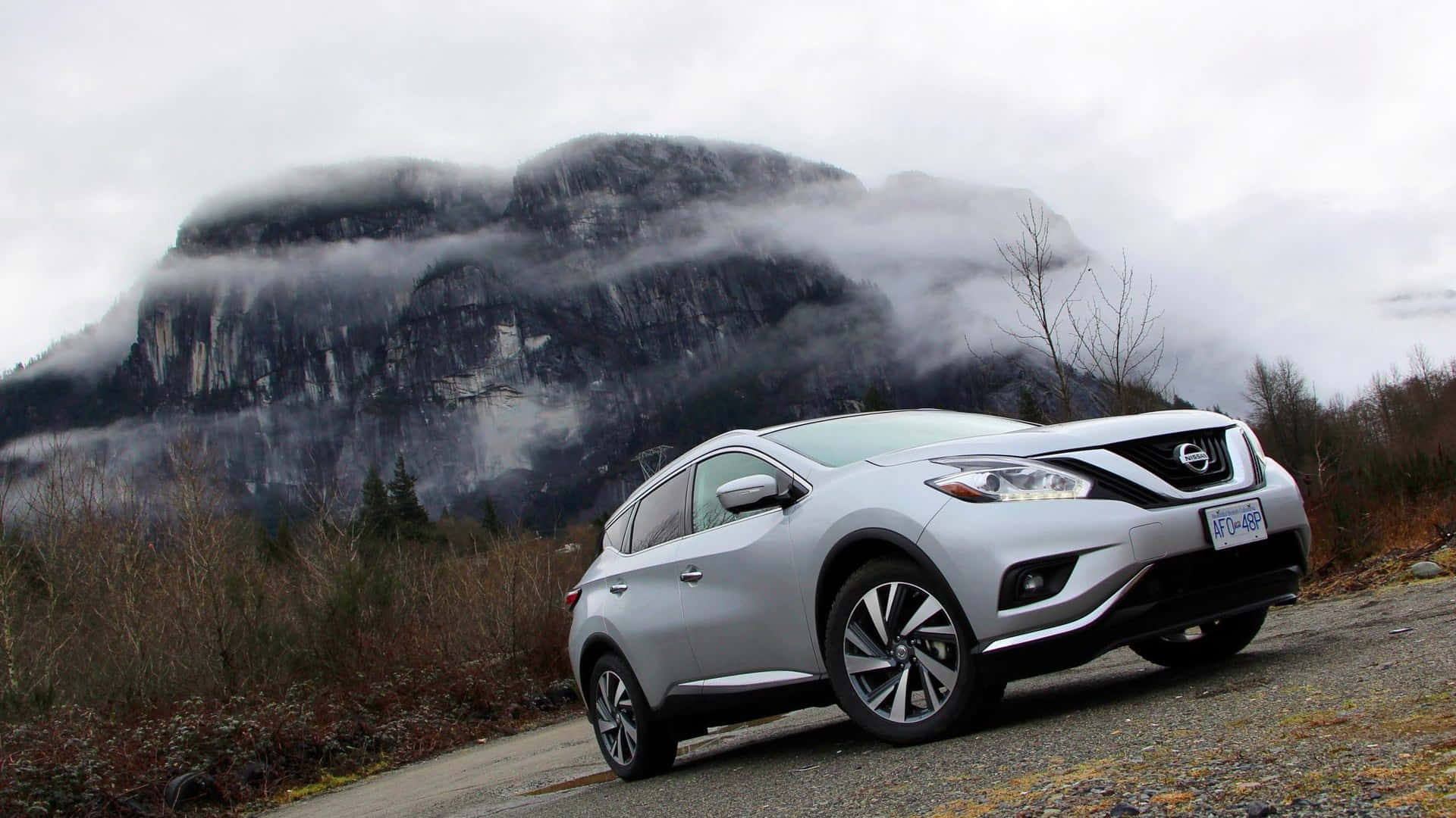 Nissan Murano 桌布 壁紙 — 一輛銀色日產Rogue SUV停泊在碎石路上，背景是霧氣瀰漫的山巒和光禿禿的樹木，天空多雲。
