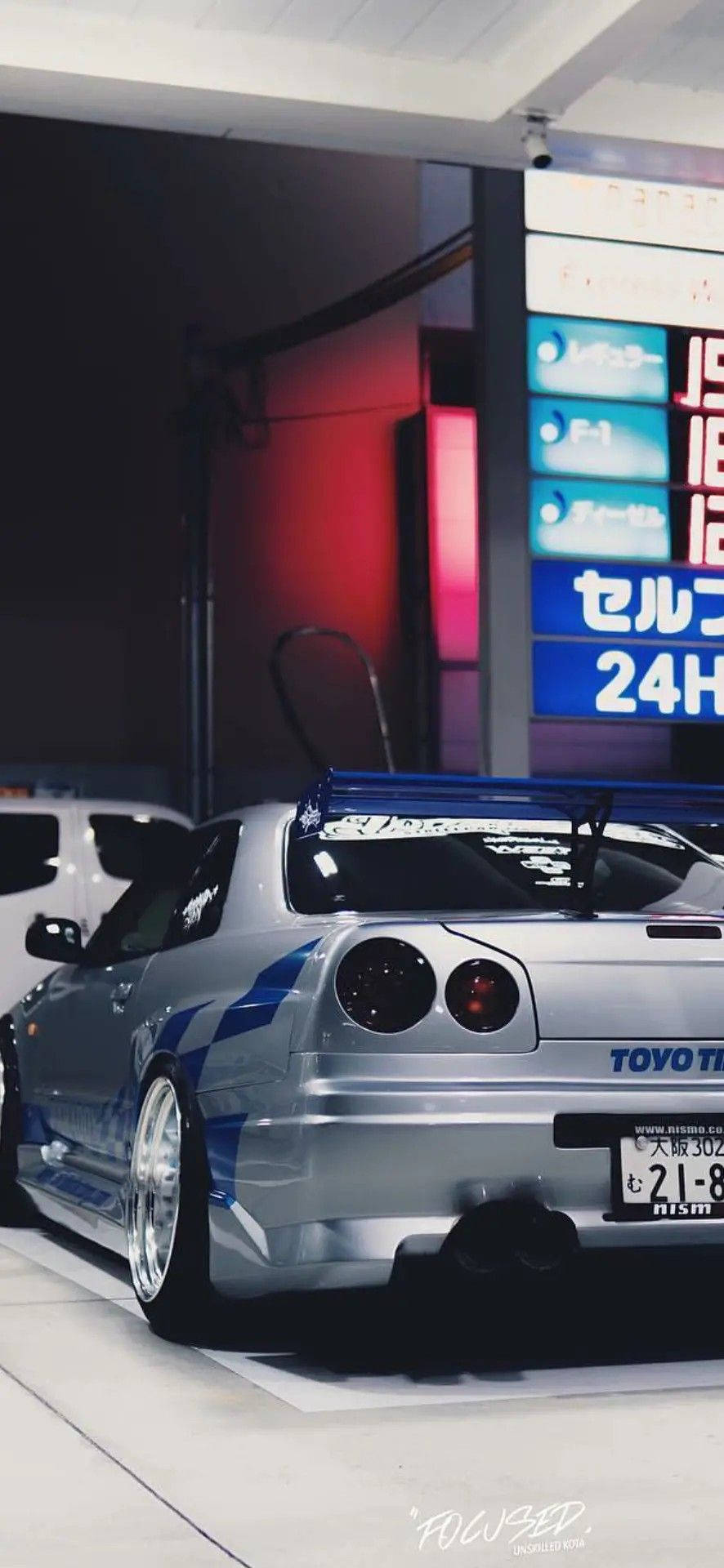 銀色日產Skyline R34帶藍色條紋，停放在發光霓虹燈招牌下。 — 保羅·沃克汽車 壁紙