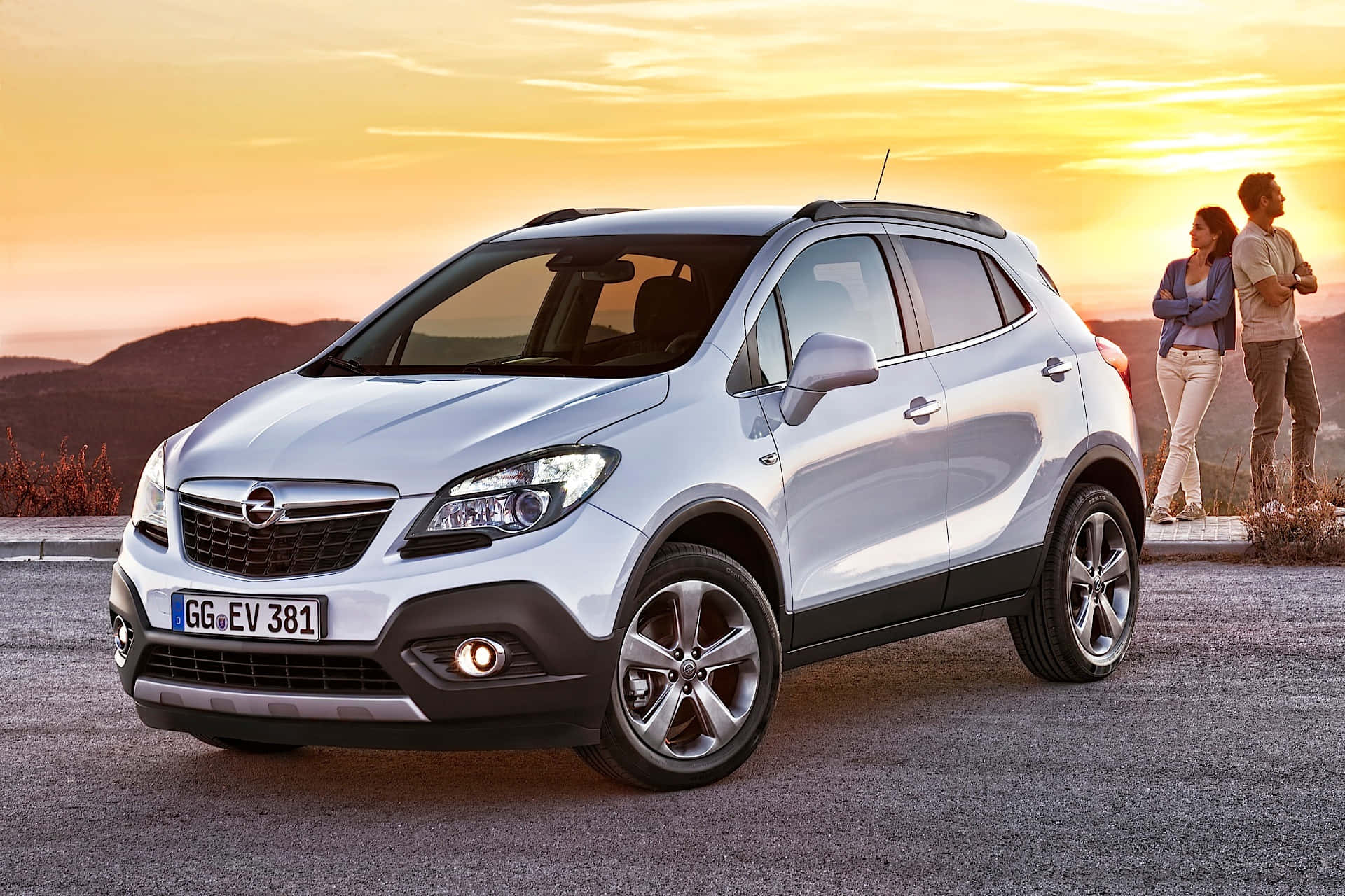 Opel Mokka 桌布 壁紙 — 一輛銀色Opel Mokka SUV停放在日落時分的碎石路上，一對情侶站在附近欣賞金色的天空。