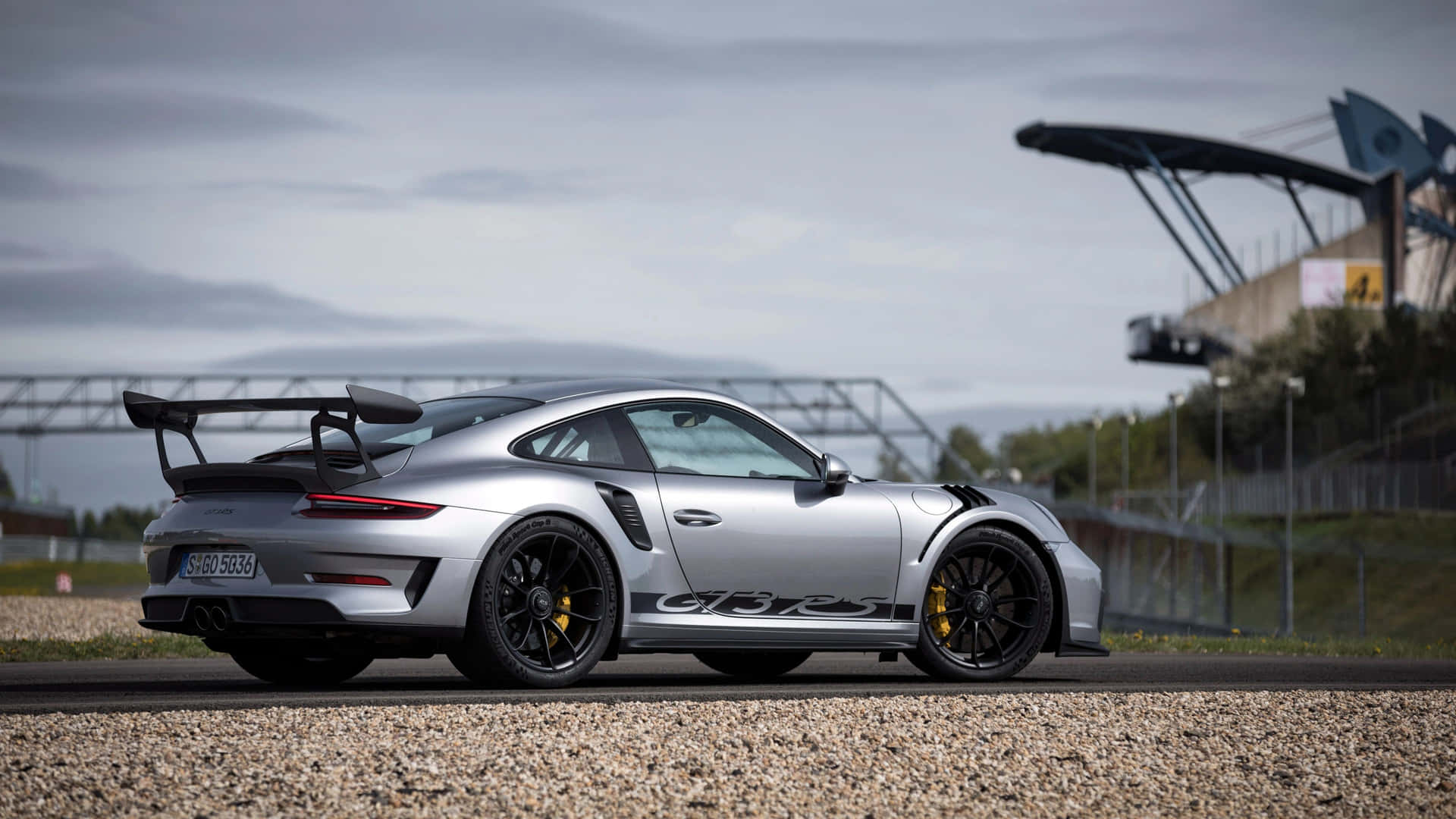 壁紙 — 一輛銀色保時捷911 GT2 RS，配備黑色輪圈和黃色制動卡鉗，停泊在賽道旁的碎石地上，天空多雲。