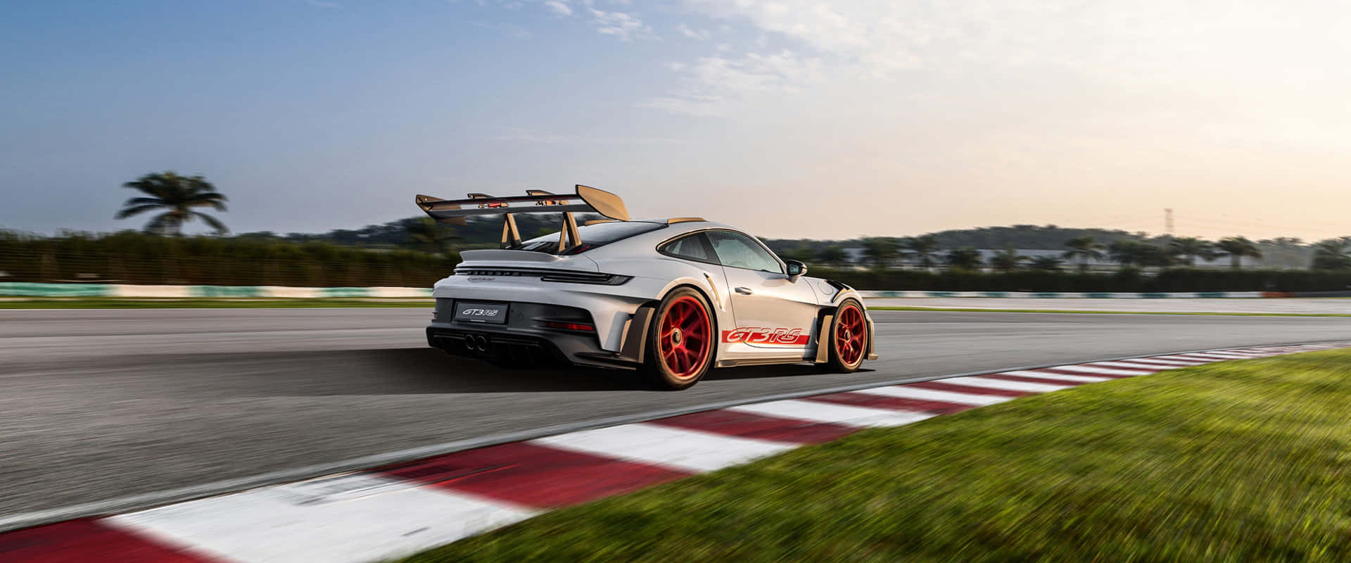  壁紙 — 一輛帶有紅色點綴的銀色保時捷 911 GT2 RS 在日落時分的賽道上奔馳，背景有棕櫚樹。