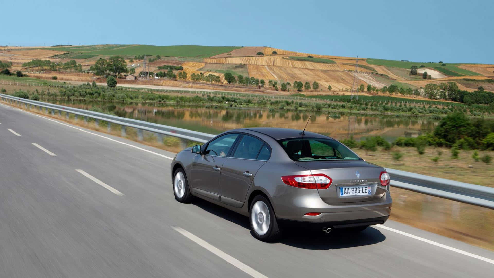 Renault Fluence 桌布 壁紙 — 一輛銀色轎車在陽光明媚的高速公路上行駛，旁邊是一條平靜的河流，背景是連綿起伏的金色山丘。