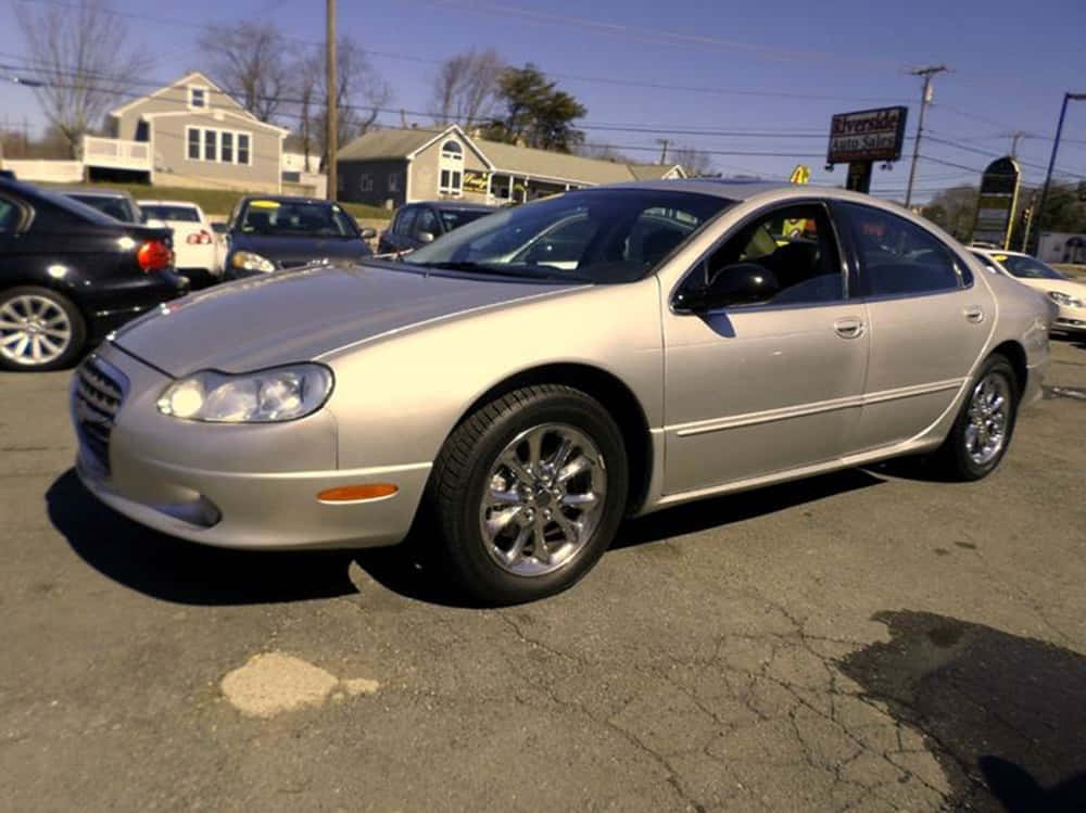 Chrysler Lhs 桌布 壁紙 — 一輛銀色轎車停放在陽光普照的二手車場，地面有裂開的柏油，背景是住宅房屋。