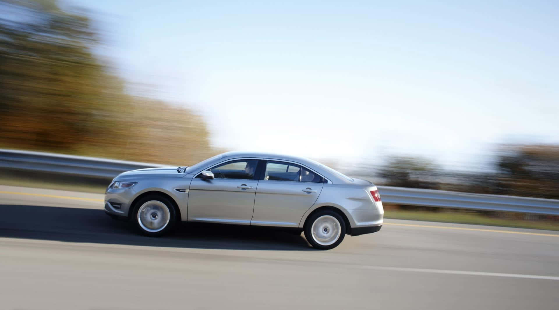 Ford Taurus 桌布 壁紙 — 一輛銀色轎車在高速公路上疾駛，背景是模糊的秋樹和護欄。