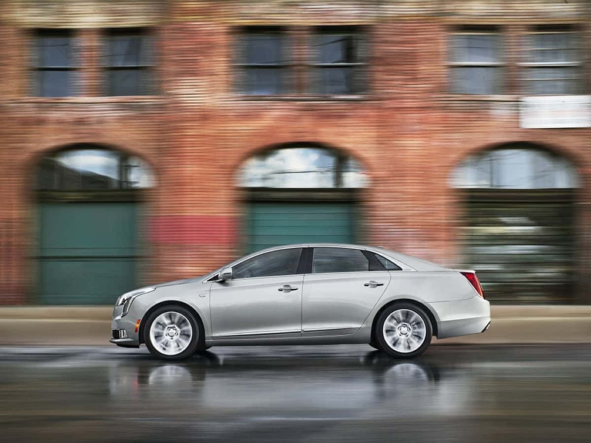 Cadillac Xts 桌布 壁紙 — 一輛銀色轎車在濕滑路面上高速駛過磚造建築，帶有動態模糊效果。