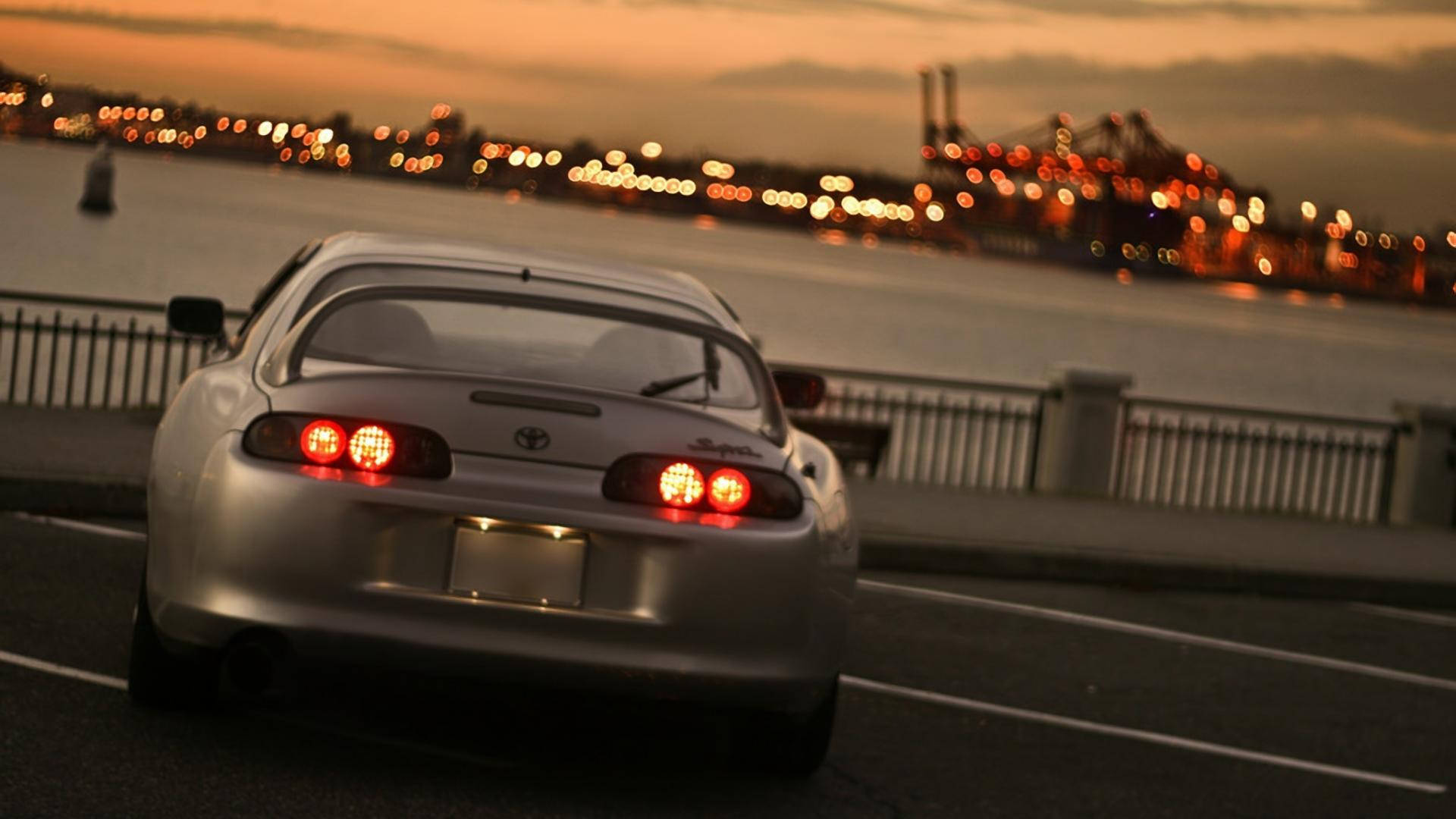 銀色跑車在黃昏時分在海濱附近行駛，背景城市燈光模糊。 — Toyota Supra 壁紙