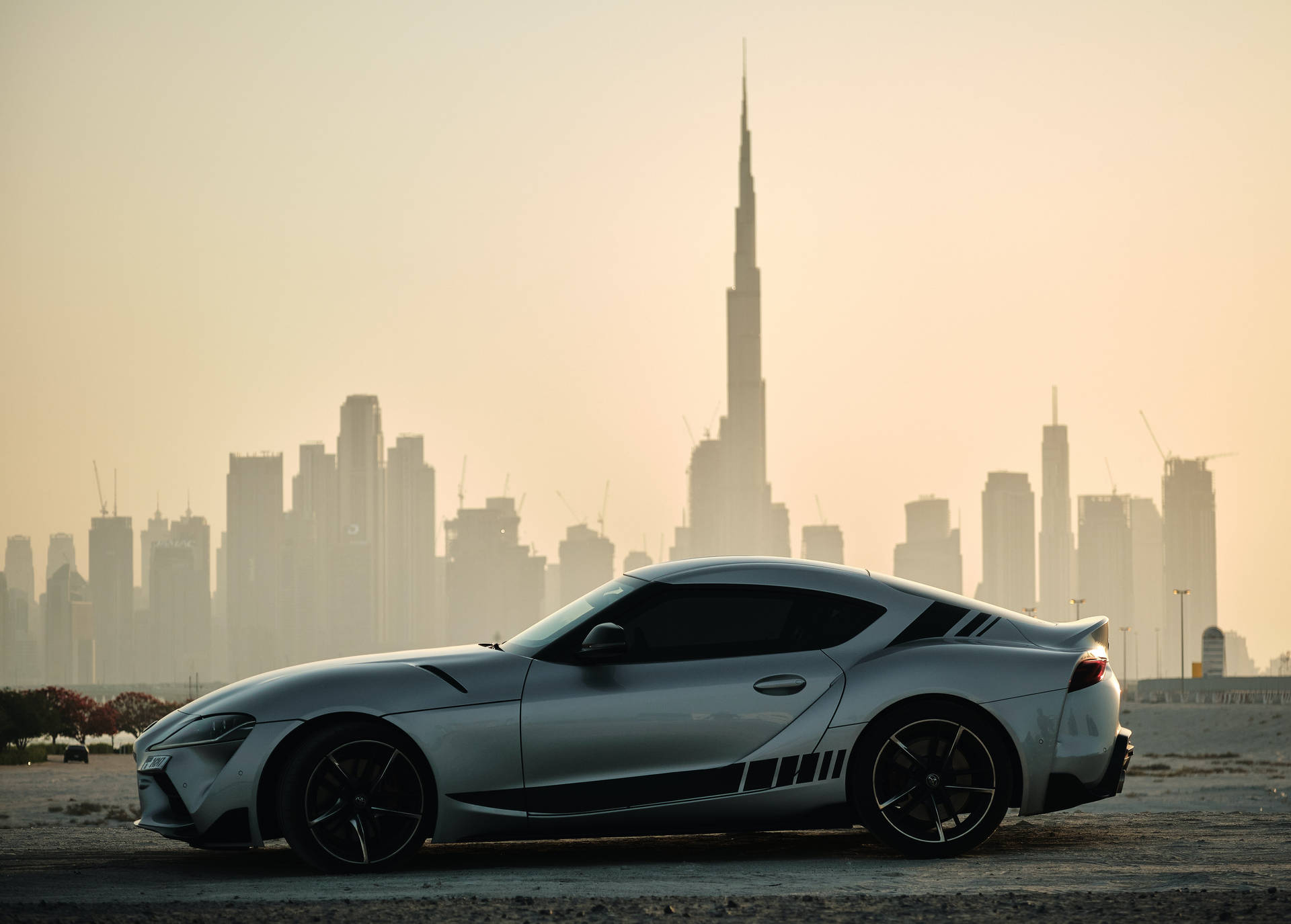 沙漠中的銀色跑車與日落時分的杜拜天際線。 — Toyota Supra 壁紙