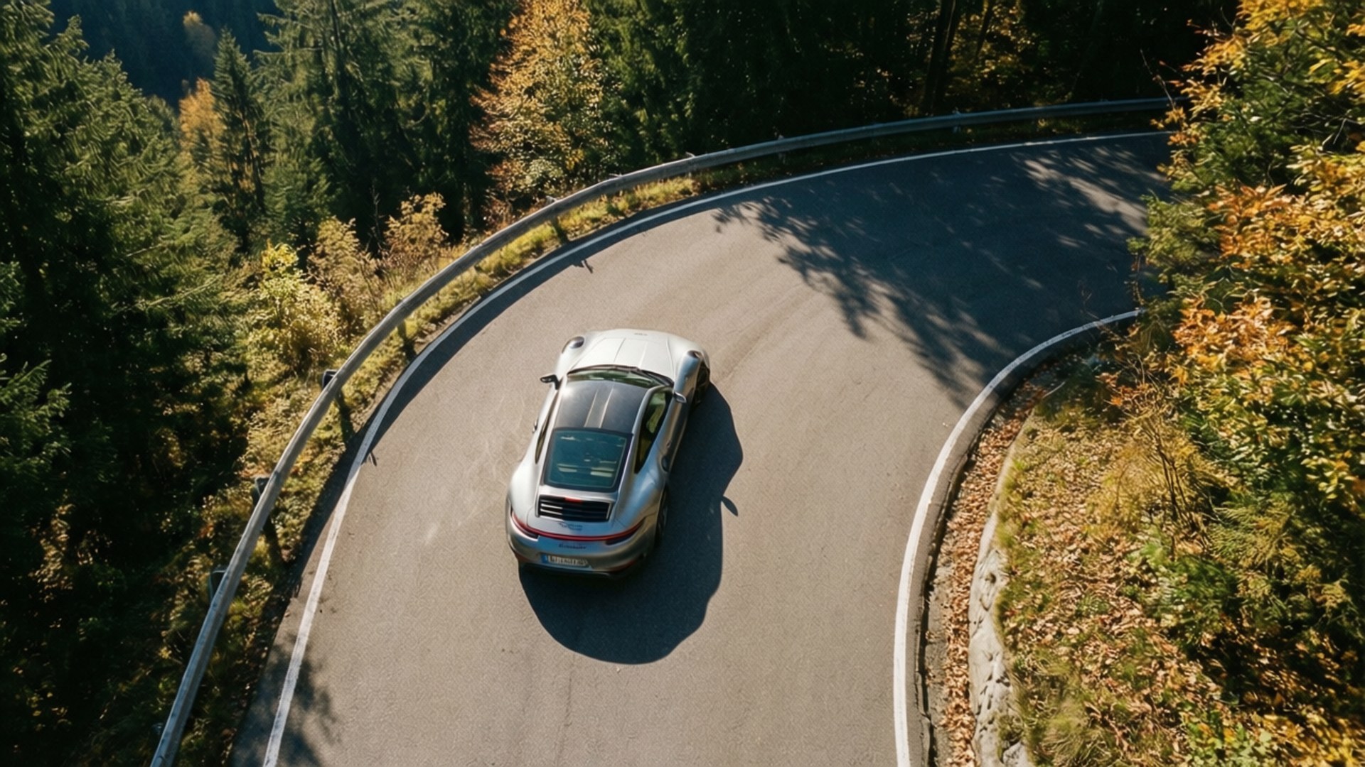 Porsche 911 Mountain Curve 壁紙 — 一輛銀色跑車在明亮的陽光下行駛在蜿蜒的山路上，兩旁是茂密的秋林。
