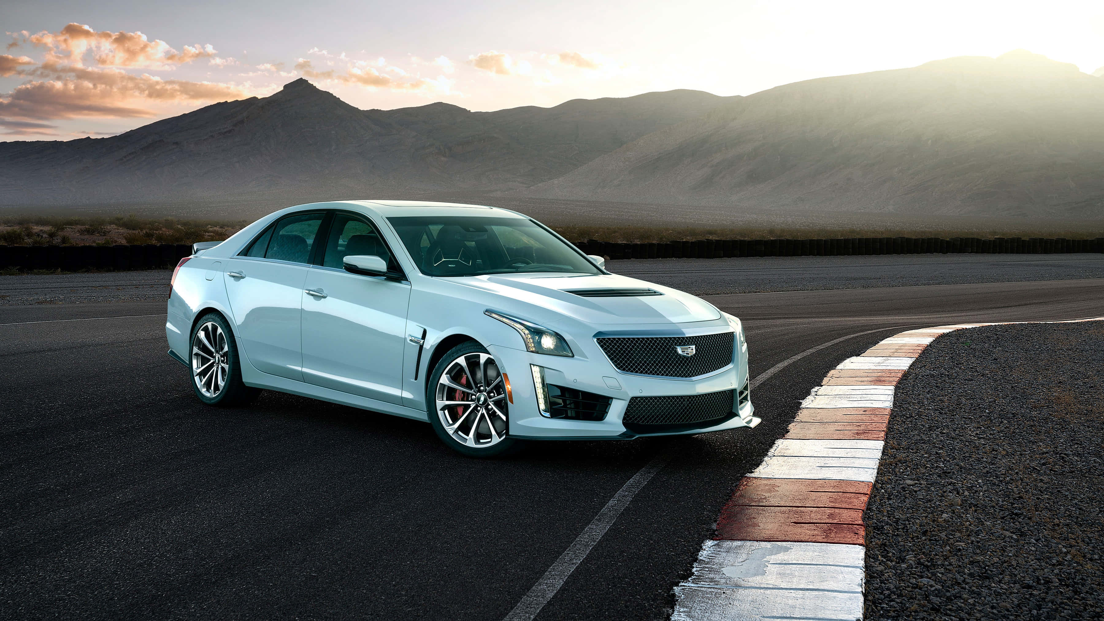 Cadillac Cts 桌布 壁紙 — 一輛流線型銀色跑車在日落時分，以群山為背景，在蜿蜒的賽道上行駛。