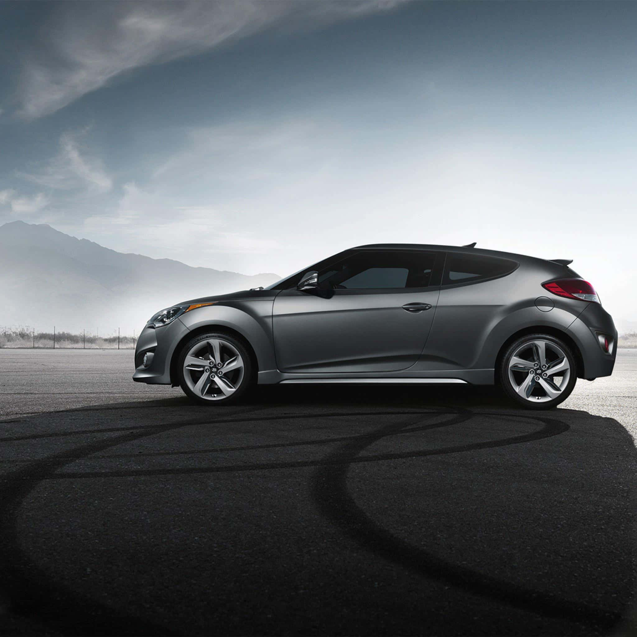 Hyundai Veloster 桌布 壁紙 — 一輛時尚的銀色跑車在瀝青路上留下輪胎痕跡，背景是霧濛濛的山脈和陰天。