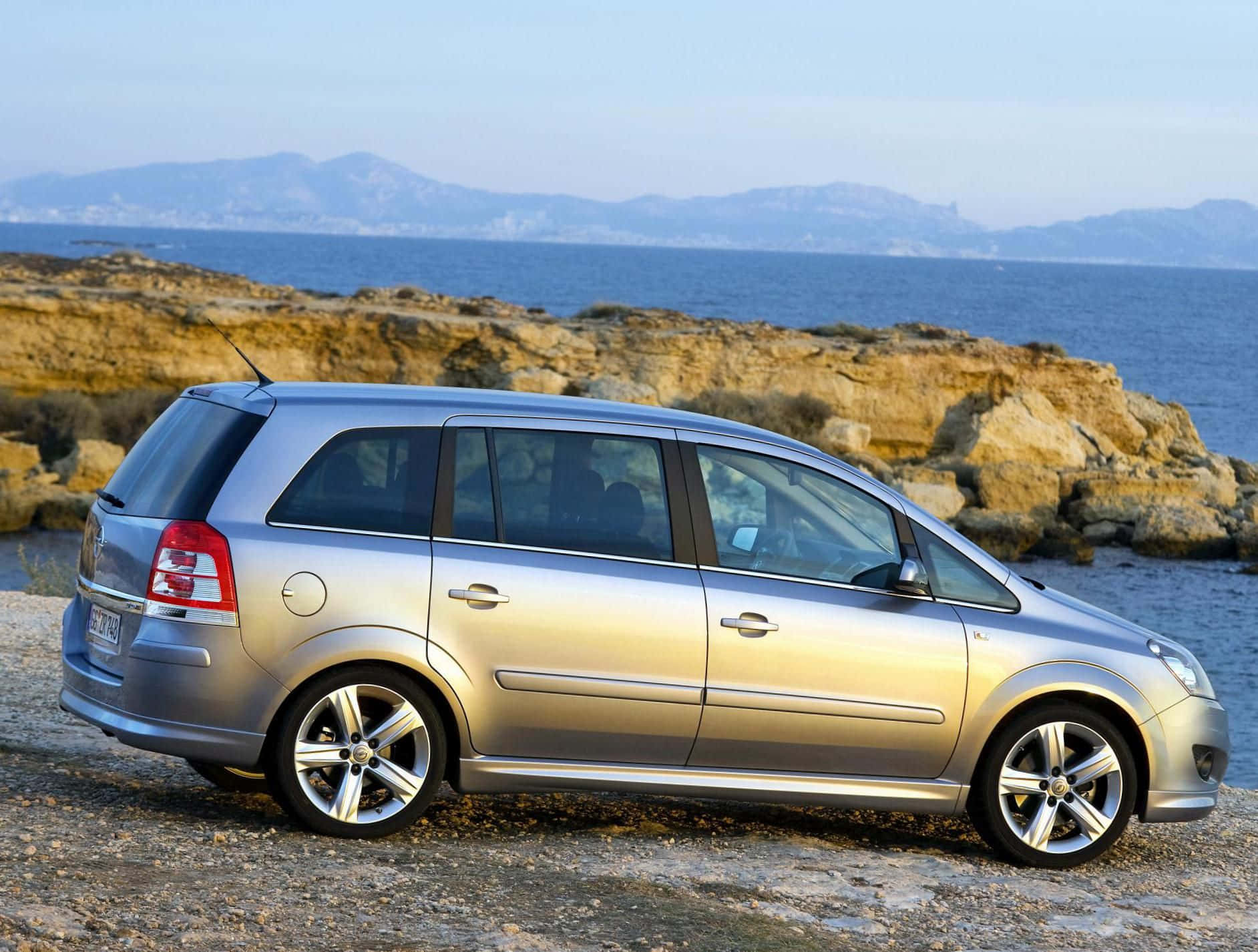 Opel Zafira 桌布 壁紙 — 一輛銀色旅行車停泊在岩石海岸上，背景是藍色大海和遠處的山脈，天空晴朗。