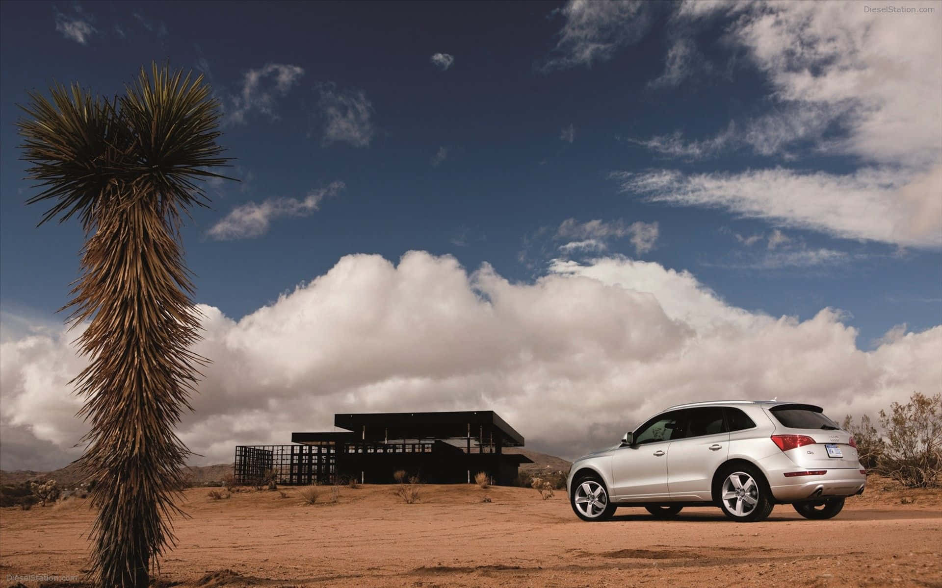 Audi Q5 桌布 壁紙 — 一輛銀色SUV停在沙漠地形上，旁邊有約書亞樹、現代黑色房屋和戲劇性的多雲天空。