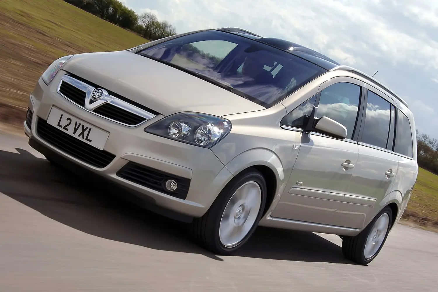 Opel Zafira 桌布 壁紙 — 一輛銀色的Vauxhall Zafira在一條有模糊草地和多雲天空作為背景的道路上行駛。